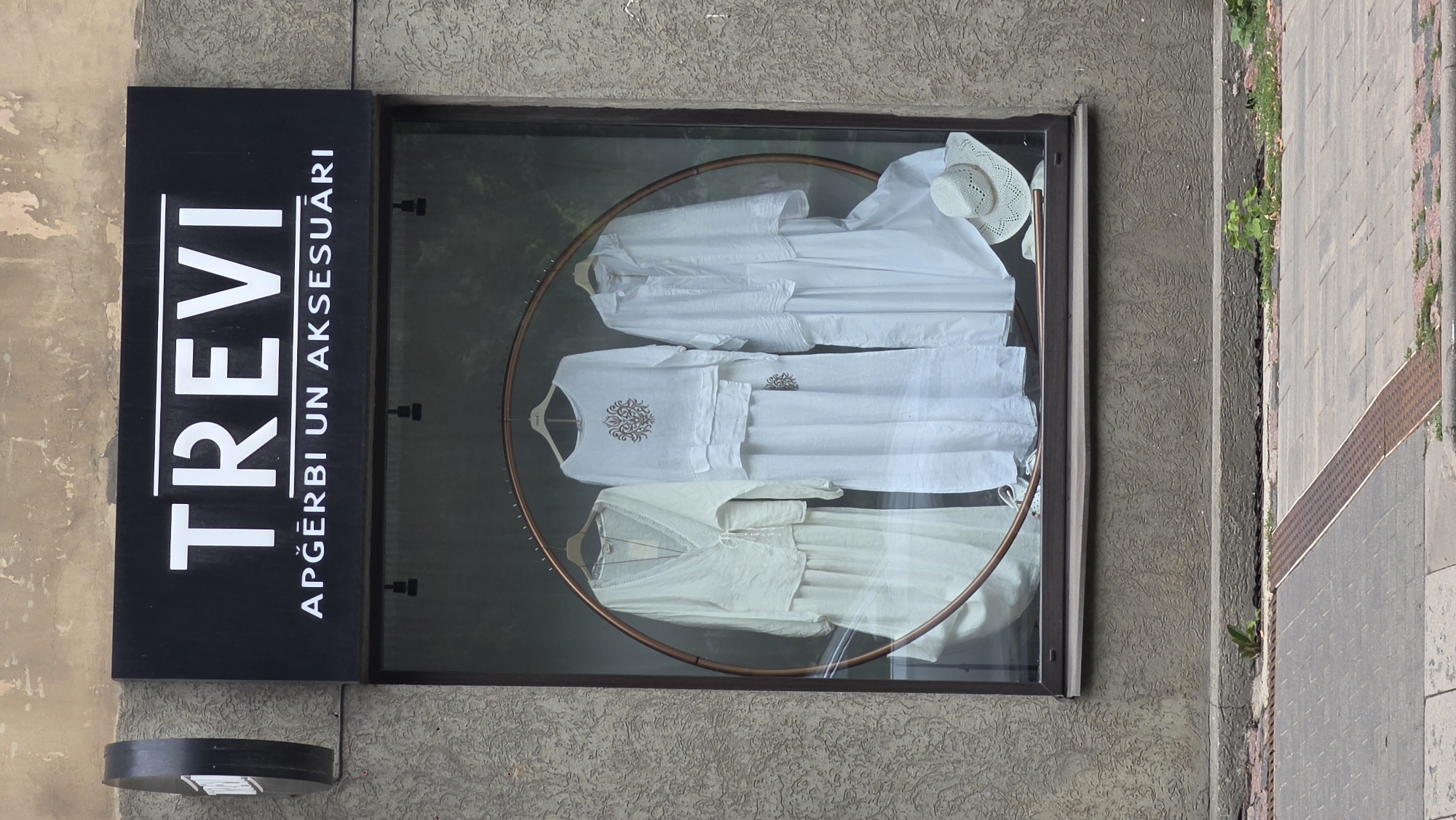 The image shows a storefront window display featuring several white linen garments and a straw hat. The garments are hanging on hangers, and the overall presentation is minimalist and elegant. The display is framed by a dark border, and behind the clothes, a circular copper-colored frame is visible. A white straw hat with a patterned brim rests on top of one of the garments.
To the left of the window is a black sign with white lettering that reads "TREVI" in large font. Below this, in a smaller font, is the text "APĢĒRBI UN AKSESUĀRI", which translates to "CLOTHING AND ACCESSORIES" in Latvian.
The building facade around the window is textured and appears to be made of concrete or stucco. To the right of the storefront, a paved sidewalk is visible, with a row of red bricks and a metal grate. Some greenery sprouts from cracks between the paving stones.
The lighting suggests it is daytime, likely overcast given the diffused light. The scene appears to be on a street in Riga, Latvia, given the text in Latvian on the sign. There are no people visible in the image, and the focus is solely on the display and the surrounding architectural elements. The overall mood is calm and clean, highlighting the fashion items.
