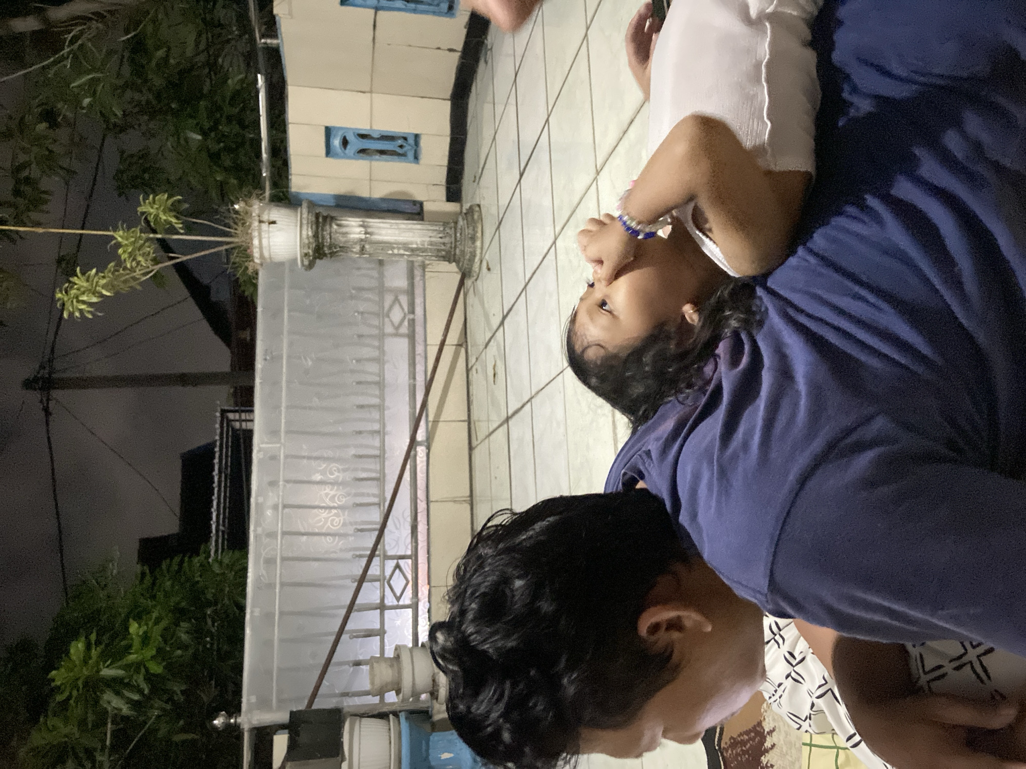 A child wearing a white shirt and blue bracelet is being held by an adult wearing a navy blue shirt. They are on a tiled surface near a building with a decorative blue accent and a potted plant. The location is possibly Gelam, Indonesia, based on the provided information.
