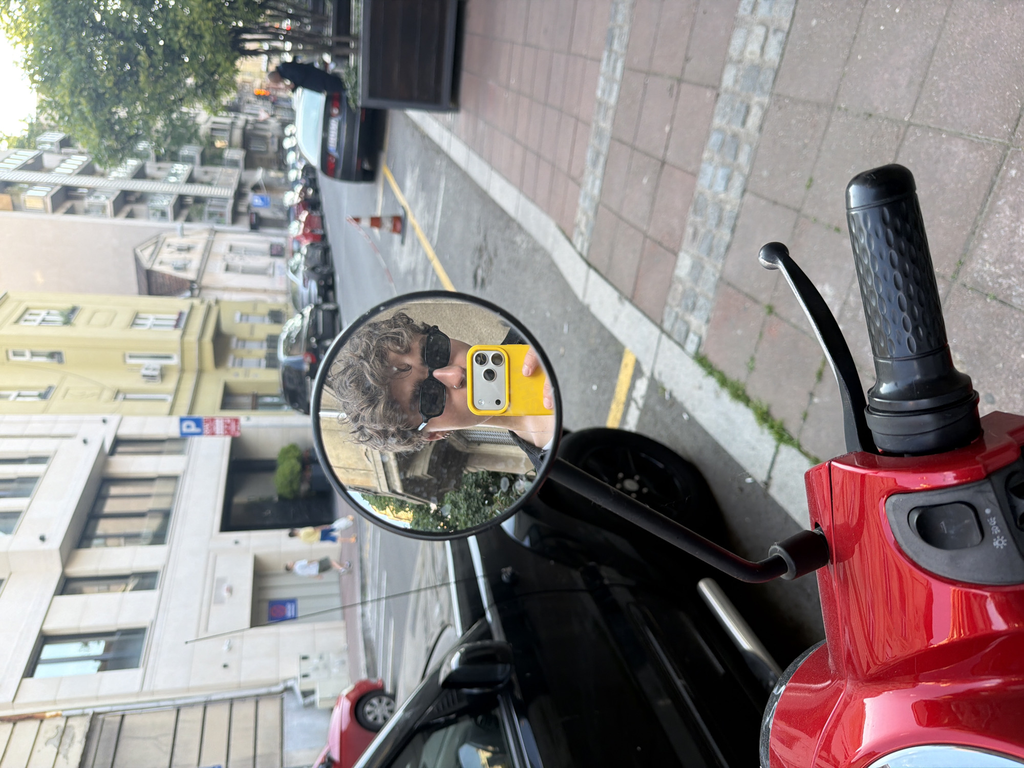 The image depicts a selfie taken in the side mirror of a red scooter. The reflection in the mirror reveals a person with curly hair, wearing sunglasses and holding a yellow smartphone. The background visible in the mirror shows a street scene, including buildings, parked cars, and pedestrians. The location is Belgrade, Serbia, as identifiable by the architecture and street layout. The scooter is parked on the side of the road. The street has a brick and paved sidewalk next to the curb. A black car is visible in front of the scooter. Further down the street are other parked vehicles.