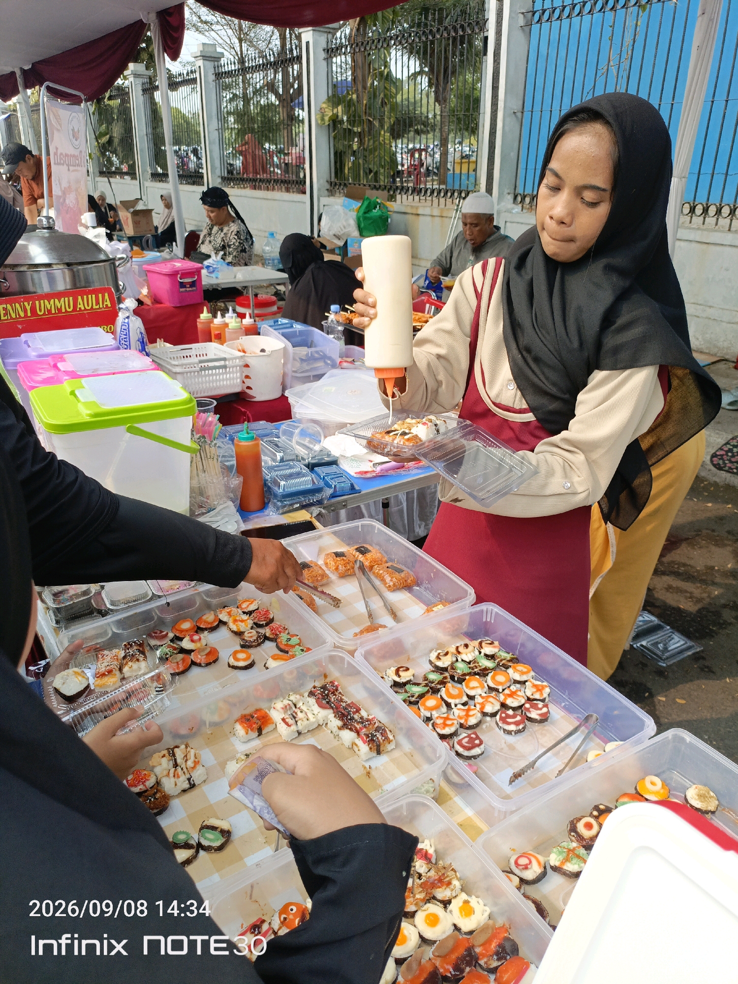 This is a medium shot of an outdoor food stall in East Jakarta, Indonesia, on a sunny day. A young woman wearing a black hijab and a maroon apron is drizzling white sauce from a bottle onto a plate of sushi rolls. She is standing behind a table laden with various food items, including more sushi rolls in plastic containers. In the foreground, a person's arm and hand, wearing a black shirt, are visible reaching towards the sushi with tongs. The stall has a red and white striped awning overhead, and a black metal fence is visible in the background, along with other people in the distance. The overall scene depicts a busy market or street food environment.