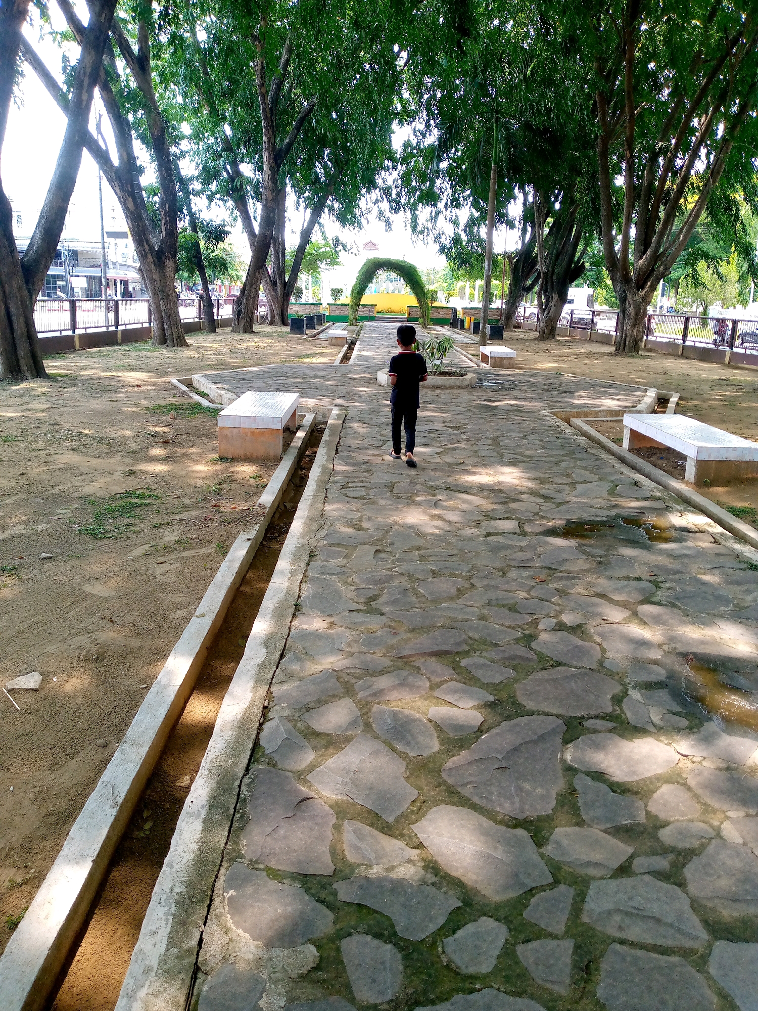 The image depicts a young boy walking on a stone-paved pathway in a park. The pathway is irregular in shape, with large, flat stones of varying sizes. To the left of the pathway is a dirt area with a concrete drainage channel. To the right of the pathway are two white benches. The park is lined with mature trees with lush green foliage, casting dappled shadows on the ground. In the background, a yellow archway with green decorative elements is visible, suggesting it might be an entrance to another area or a landmark within the park. The overall environment appears to be a public park in Lhokseumawe, Indonesia, possibly on a sunny day, given the shadows.