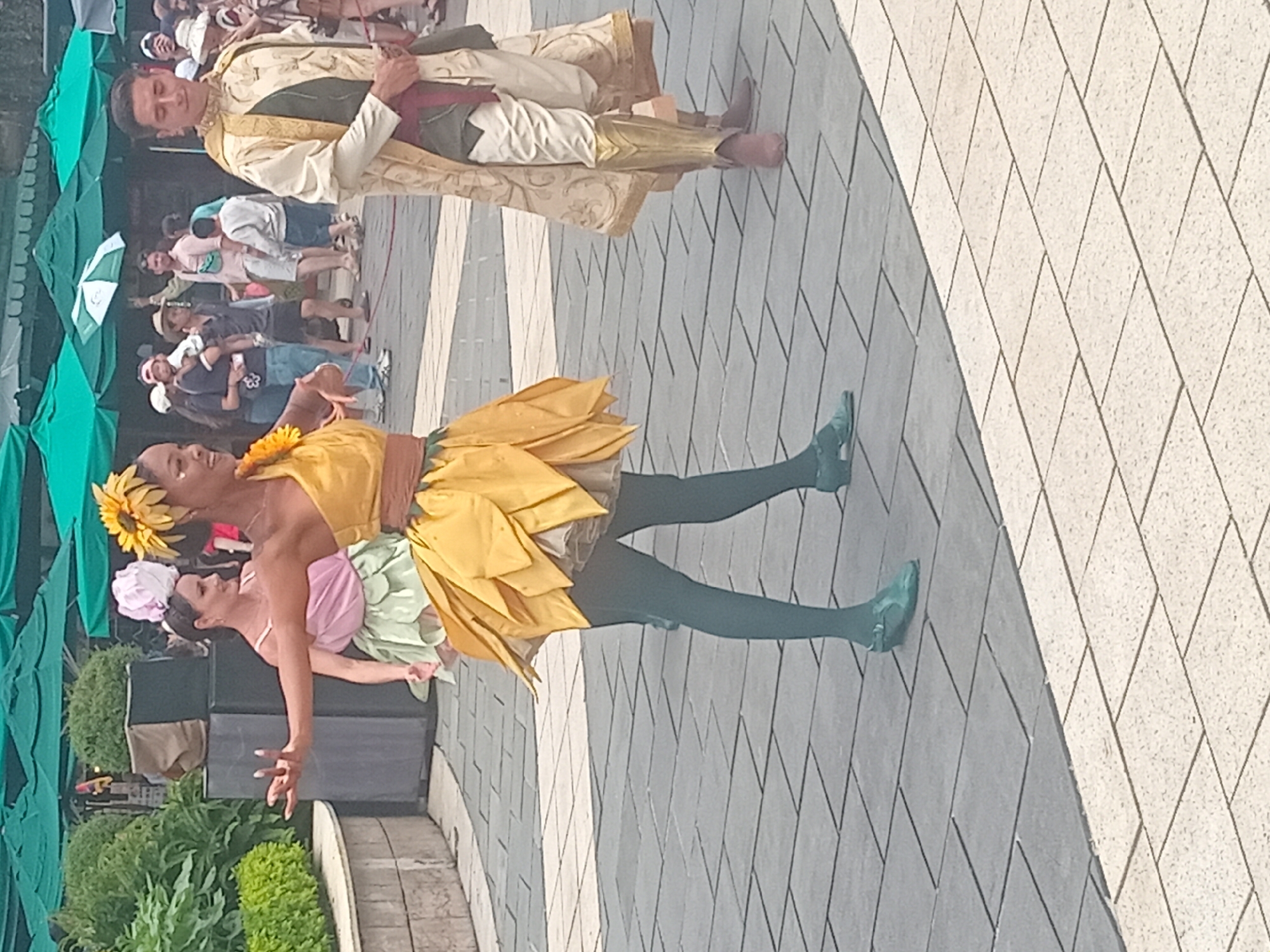 A group of people are gathered outdoors in what appears to be a plaza or walkway. In the foreground, a woman in a yellow, petal-like dress and a sunflower hair accessory is dancing with her arms outstretched. To her right, a man in a gold and cream colored robe stands with his hands clasped. Further back, another woman in a pink and green outfit can be seen. The ground is paved with gray and light-colored tiles. In the background, there are green umbrellas, lush greenery, and other onlookers. The scene suggests an outdoor event or performance.