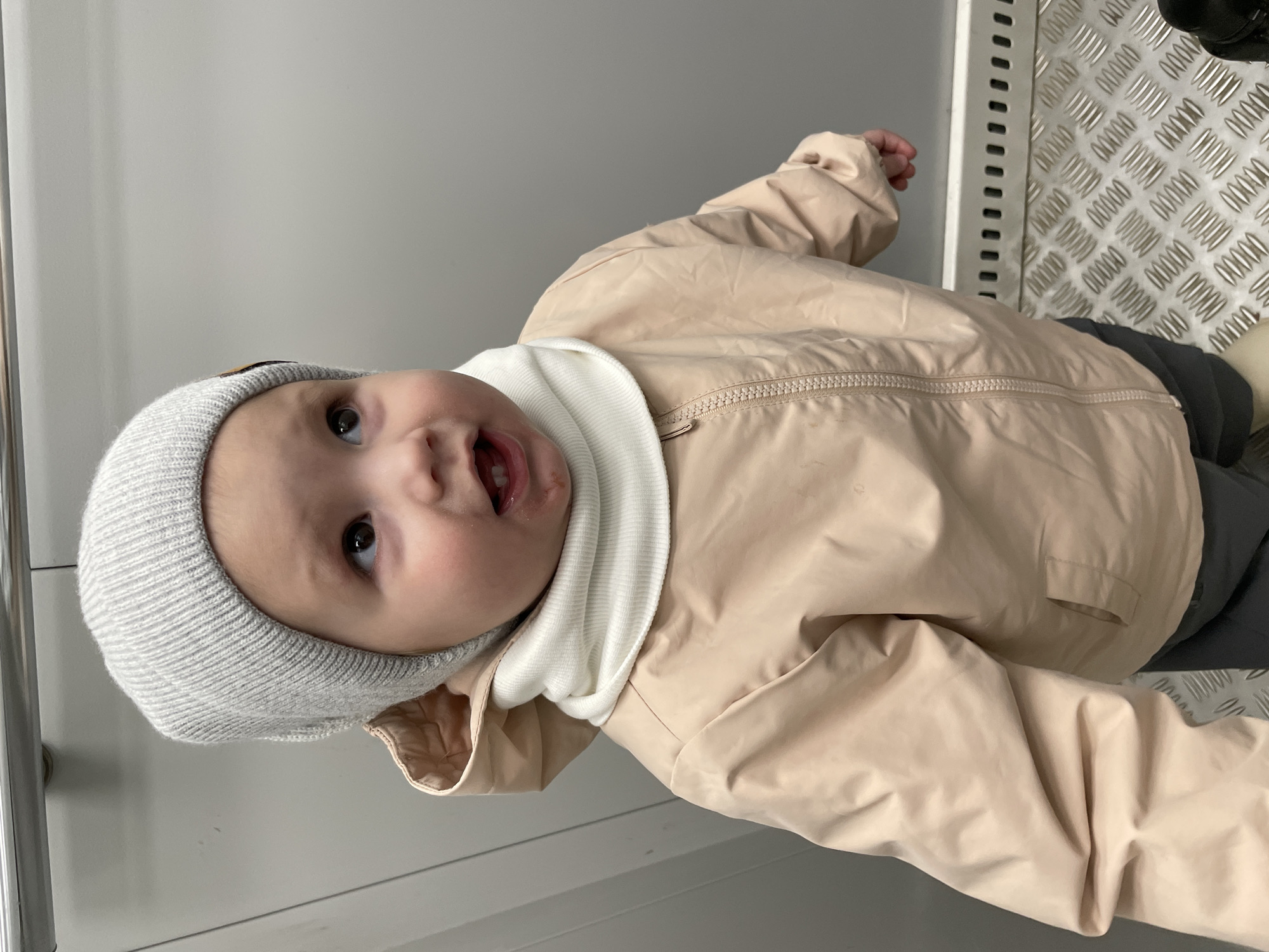 A baby wearing a gray knitted hat, a white turtleneck, and a beige jacket is lying on a gray surface. The baby is looking upwards with an open mouth. To the right of the baby is a diamond-patterned metal surface next to a white vertical panel. To the left of the baby is a vertical silver pole and a white surface. The location is not discernible as Izhevsk, Russia.