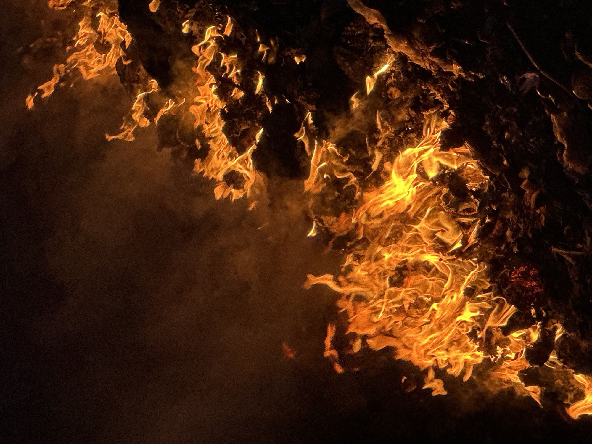 The image depicts a close-up view of a fire. Flames of orange and yellow consume what appears to be a solid, possibly organic material. There is heavy smoke and a dark background, suggesting a nighttime setting. The location cannot be determined from the image. There are no visible people.