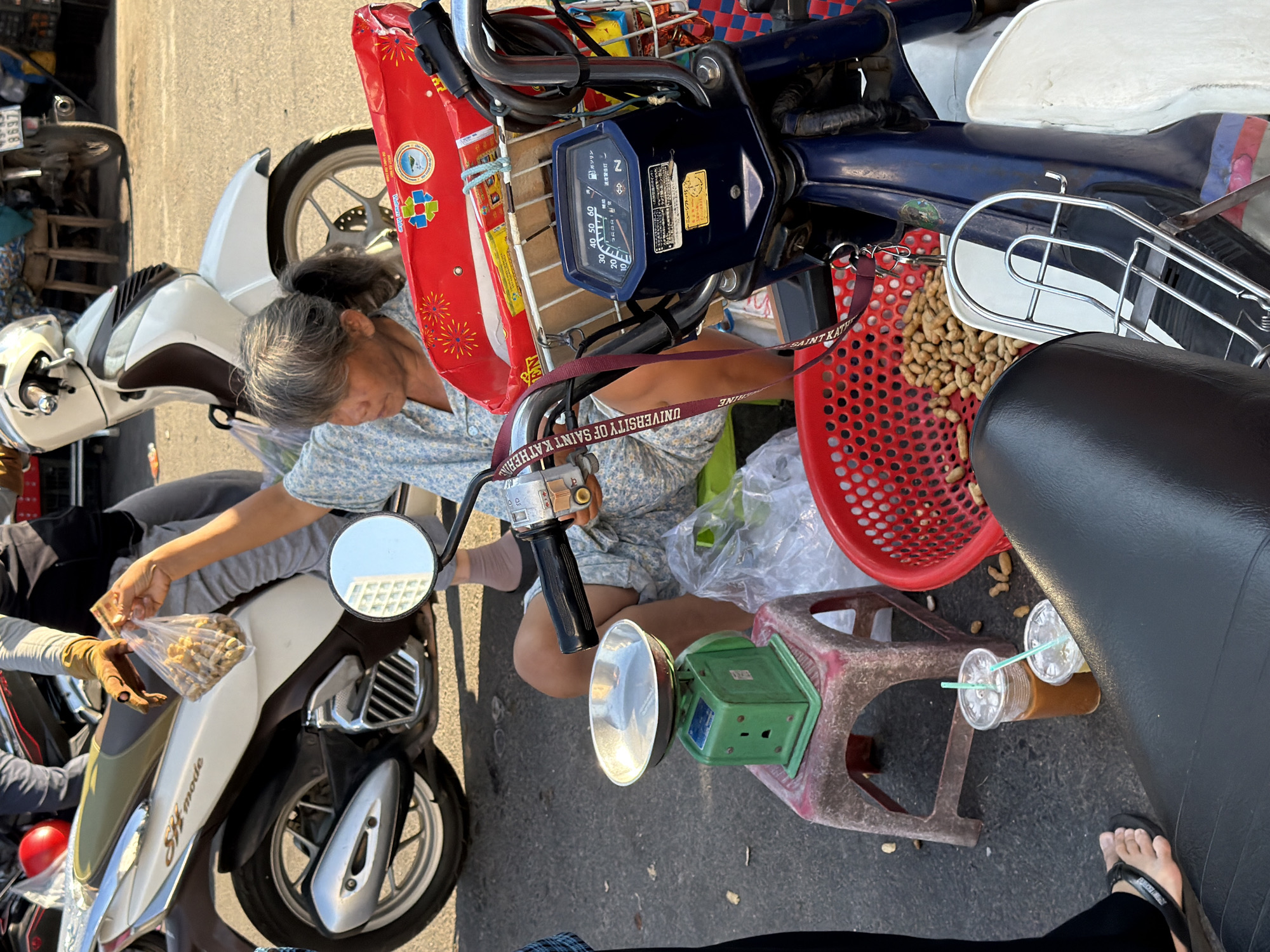 A woman with gray hair pulled back in a ponytail, wearing a blue and white patterned shirt, sits on a motorcycle in what appears to be a street market in Nha Trang, Vietnam. She is holding a clear plastic bag filled with small, light-brown, shelled nuts, offering them to someone out of frame. In front of her, a green mechanical scale sits on a red and white plastic stool. Beside the scale are two plastic cups with straws, one containing a dark brown beverage and the other a lighter brown liquid. A red plastic basket overflowing with similar nuts is located to the right of the stool. The background shows other motorcycles, one of which is white and cream-colored. The ground is paved and appears to be asphalt.