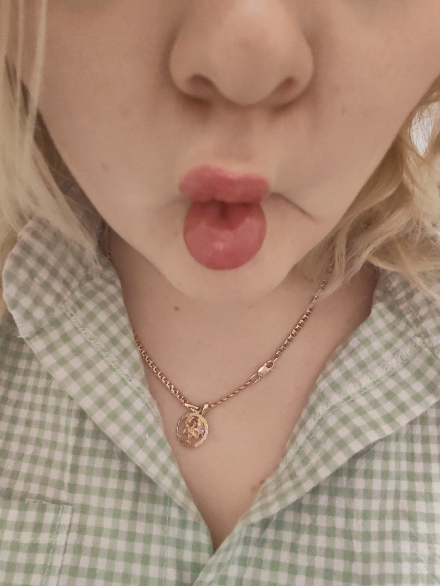 A close-up shot shows a person from the nose down to the upper chest. The person has fair skin and blonde, wavy hair. Their lips are pursed into a "fish face" expression. The person is wearing a green and white gingham shirt. A gold chain necklace with a round pendant hangs around their neck. The background is out of focus and indistinct. There is no indication of the city of Izhevsk, Russia.