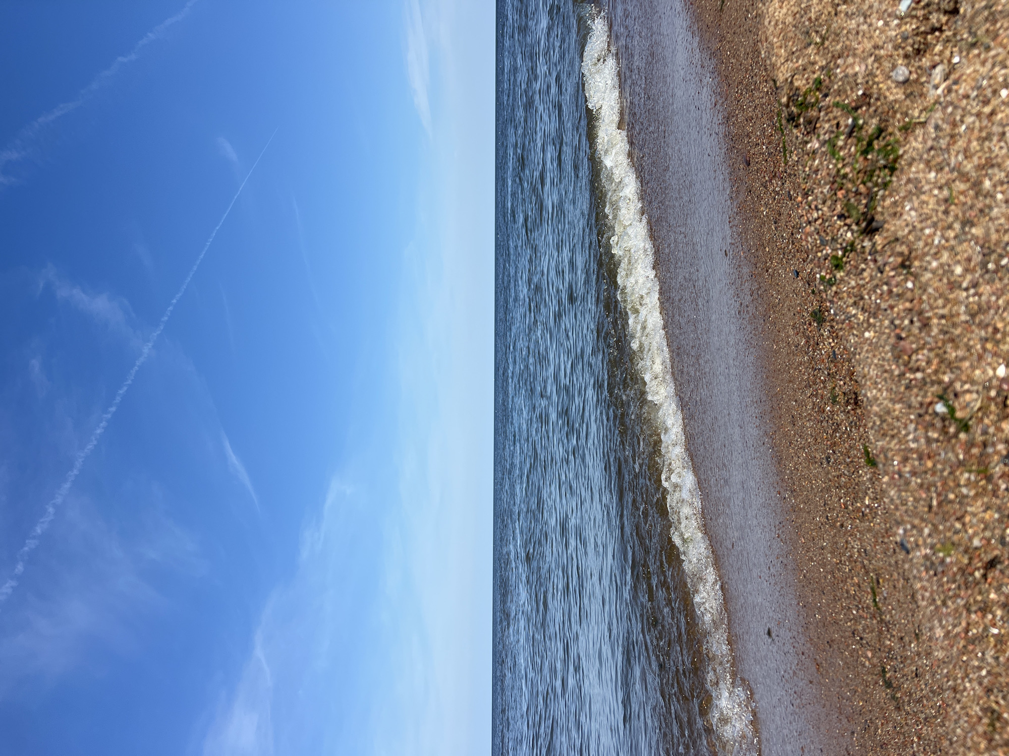 A low-angle, wide shot captures a tranquil scene at the edge of a body of water. The foreground is dominated by a sandy beach composed of small pebbles and grains in varying shades of brown, with a few patches of green algae. A gentle wave is breaking onto the shore, its white foam creating a delicate line of effervescence. The water itself is a deep blue, with subtle ripples indicating a light breeze. In the distance, the horizon is a sharp, straight line where the water meets the sky. The sky is a clear, vibrant blue, with thin, wispy white clouds scattered across it, including two prominent contrails forming an 'X' shape high above. The image is well-lit, suggesting it was taken during daylight hours.