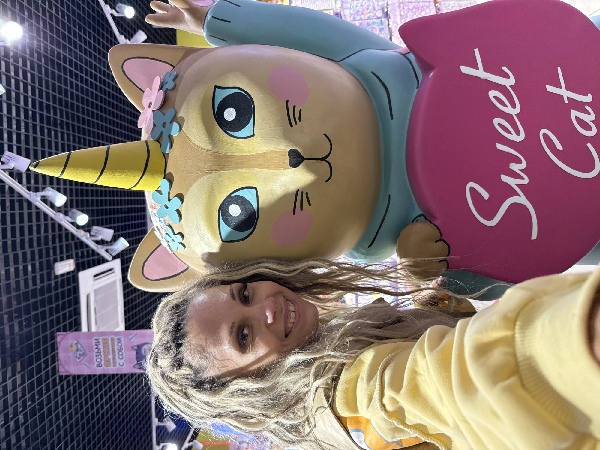 This is a medium shot of a woman with blonde dreadlocks and a yellow hoodie smiling at the camera. She is standing next to a large, cartoonish cat statue with a yellow unicorn horn and blue eyes. The statue is wearing a light blue shirt and holding a pink heart-shaped sign that says "Sweet Cat" in white lettering. The background appears to be an indoor setting, possibly a toy store or amusement park, with bright lighting and displays visible. A banner with Russian text hangs in the background. The overall scene is playful and cheerful. The location is the "Sweet Cat" store in Yaroslavl, Russia.