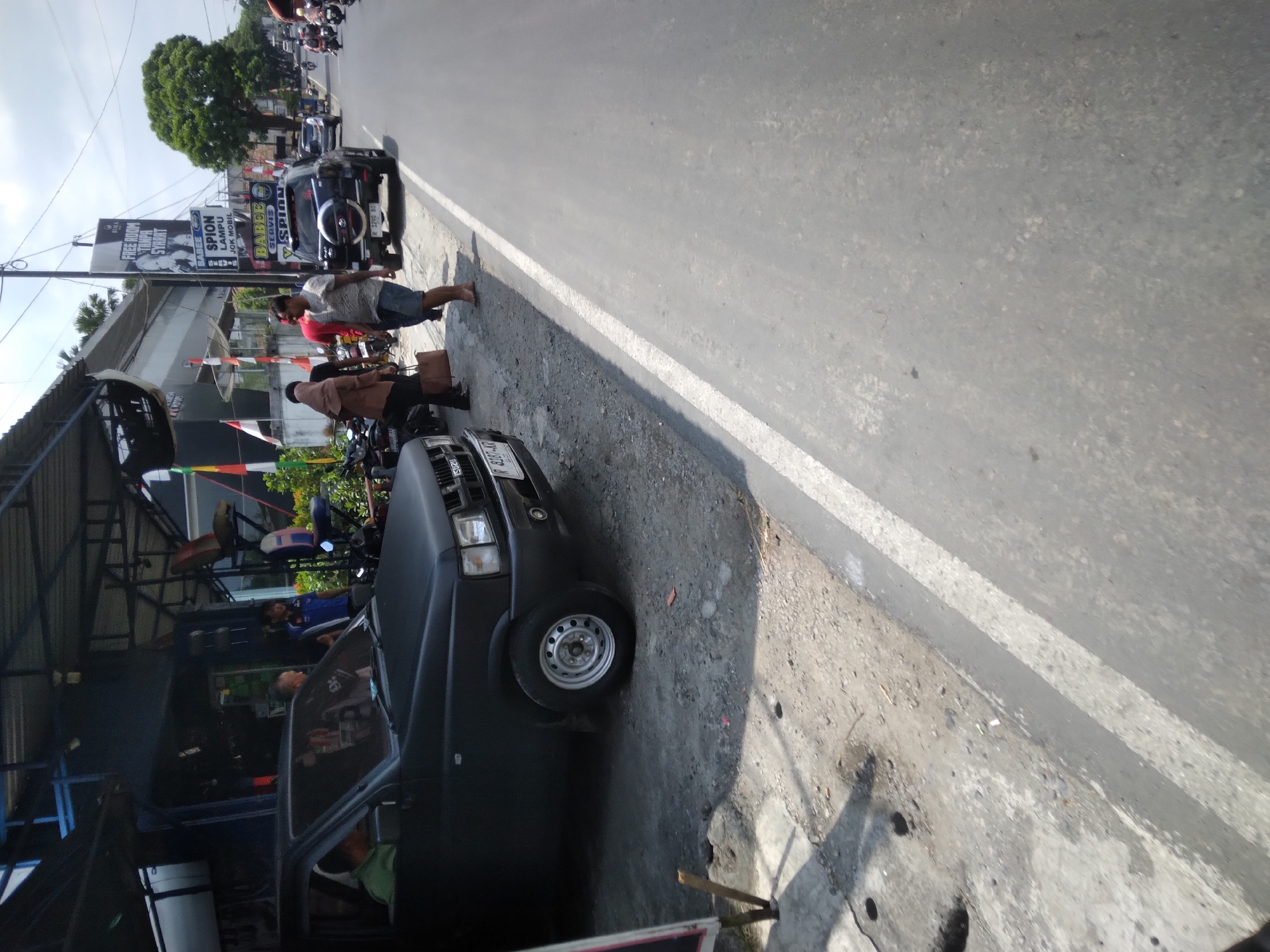The image depicts a street scene in Sokaraja, Indonesia, on a sunny day. A dark-colored pickup truck is parked on the side of the road, partially under an awning. Several people are standing and interacting near the truck and in front of a building. To the right of the truck, a black SUV with a spare tire on the back is also parked. The road is paved, with a white line marking the lane. In the background, other vehicles, including motorcycles, are visible on the road, suggesting moderate traffic. Buildings and trees line the street. A tall sign with text in Indonesian is mounted on a pole. The overall environment appears to be a commercial or roadside area.