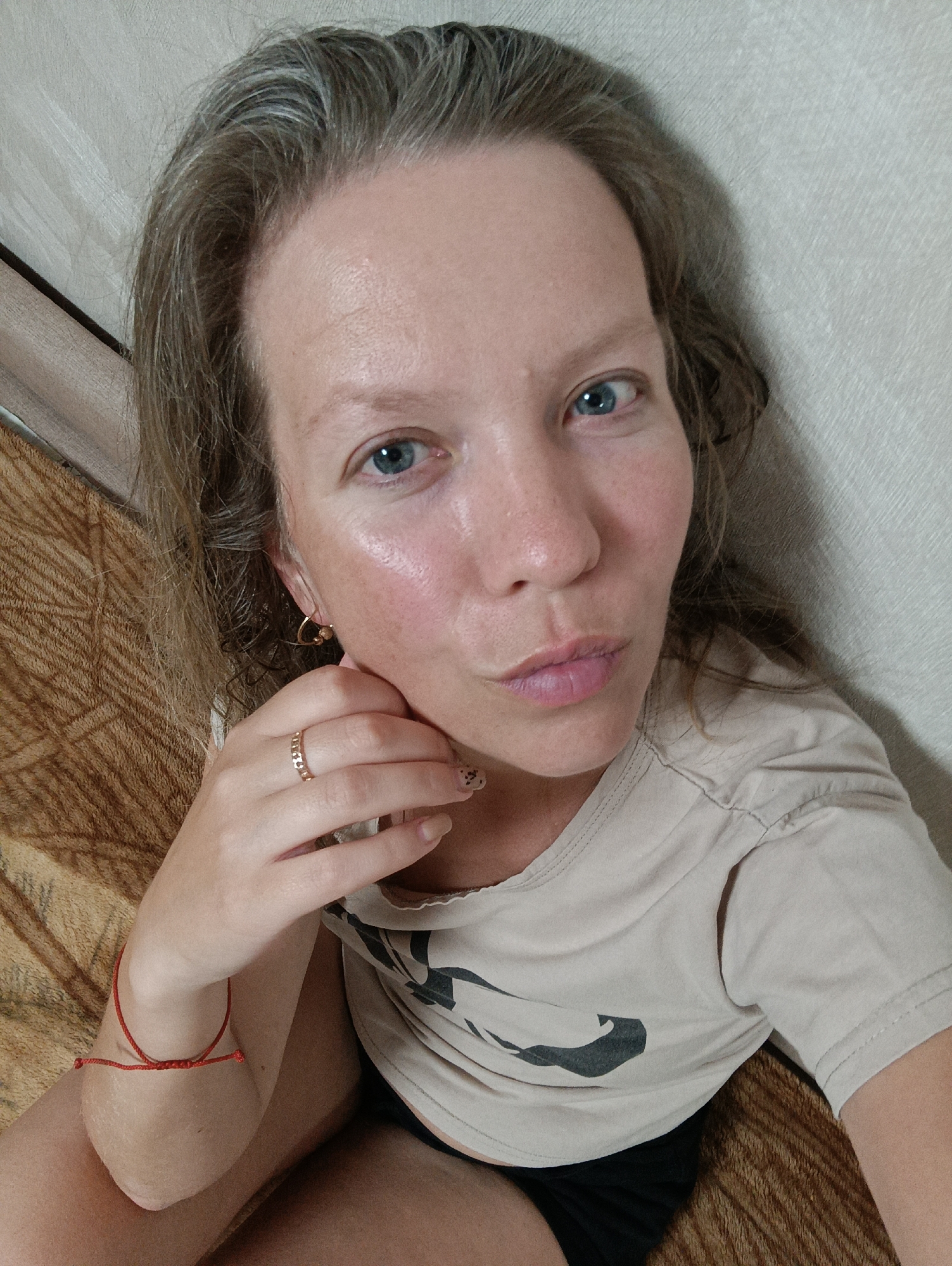 A Caucasian woman with grey-streaked brown hair and blue eyes is seen from a high-angle, close-up perspective. She is wearing a light-colored t-shirt with a graphic design on it and black shorts. A gold ring is on her left ring finger and gold heart-shaped earrings are visible. A red string bracelet adorns her left wrist. She is touching her face with her left hand. The background consists of a patterned brown blanket and a light-colored wall. The image does not provide enough information to definitively confirm the location in Chaikovsky, Russia.
