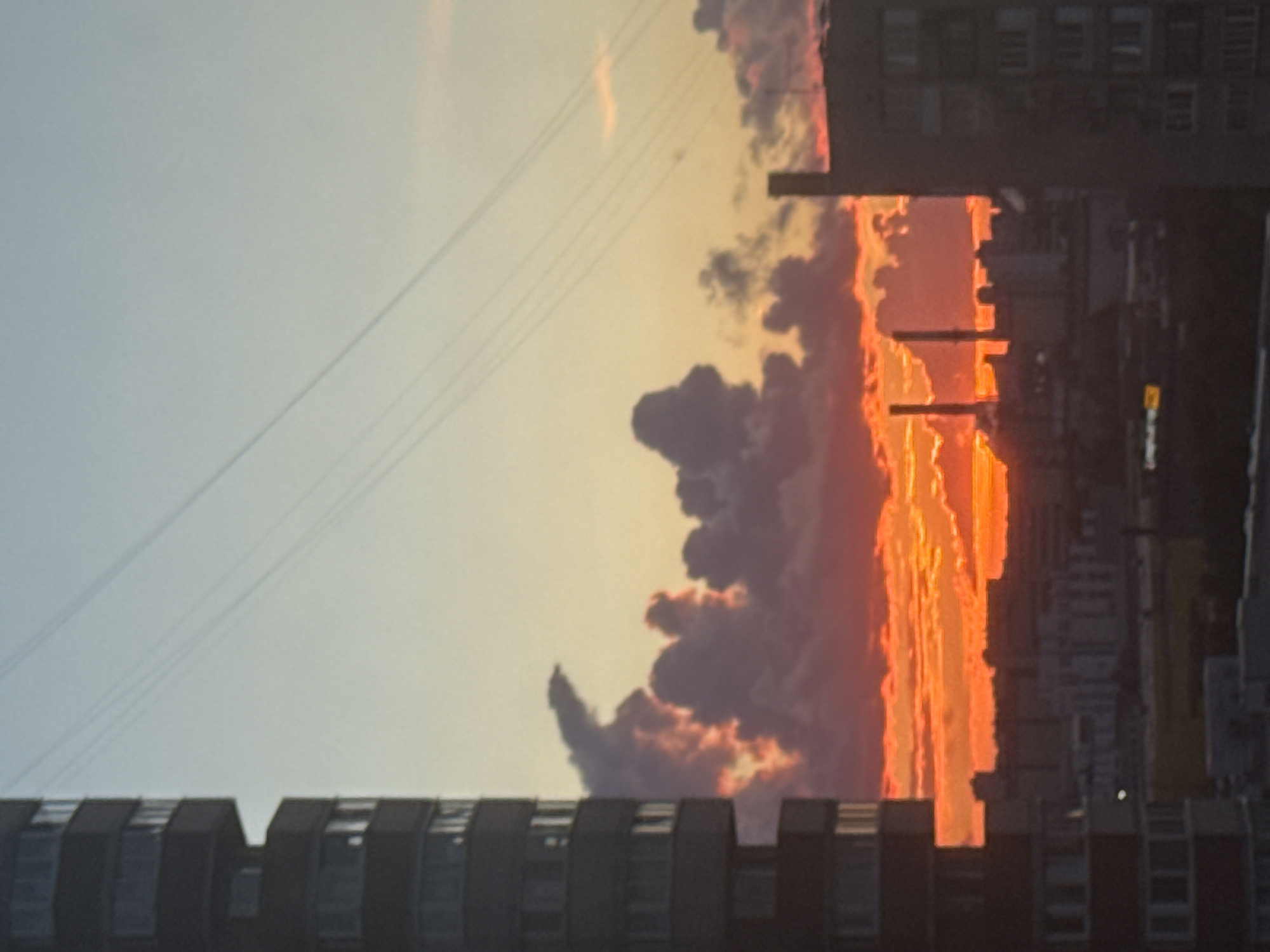 The image captures a striking sunset over the city of Saint Petersburg, Russia, with a strong emphasis on the dramatic interplay of light and shadow on the urban landscape and the sky. In the foreground, a tall, dark residential building dominates the left side of the frame, its many windows reflecting the warm hues of the setting sun. The building is characterized by its repetitive, modular design, typical of Soviet-era architecture.
The sky is a canvas of vibrant oranges, reds, and yellows, particularly concentrated along the horizon where the sun is setting. This intense color is a result of the low angle of the sun, illuminating the undersides of the clouds. The clouds themselves are a mix of dark, silhouetted masses and brightly lit edges, creating a textured and dynamic ceiling to the cityscape. Some of the cloud formations appear anthropomorphic or animalistic, adding an element of visual interest.
In the middle ground, the silhouette of the city unfolds, revealing more buildings and industrial elements. Two prominent smokestacks, characteristic of a power plant or industrial facility, rise above the lower structures. Their dark forms contrast sharply with the bright sky. The city appears dense, with numerous apartment blocks and other structures stretching towards the horizon.
Several power lines are visible in the upper portion of the image, stretching diagonally across the sky, adding a layer of human infrastructure to the natural spectacle of the sunset. These lines are thin and dark against the lighter sky.
On the right side of the image, another large residential building is partially visible, its dark facade and windows providing a counterpoint to the building on the left. The overall impression is one of a bustling urban environment bathed in the ephemeral beauty of a sunset. The image is devoid of any visible people or discernible actions, focusing solely on the atmospheric conditions and the built environment. The photograph appears to have been taken from an elevated vantage point, likely from another building, looking out over the city. The quality of the image suggests it was taken with a mobile device, with some slight motion blur and noise present.