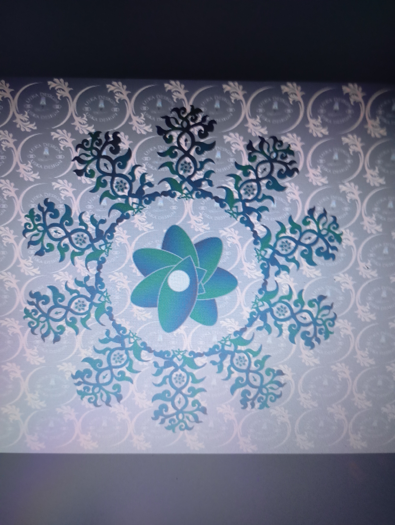 The image depicts a blue and green floral design with a central atom-like figure. The design is surrounded by a circular pattern of stylized floral elements, all set against a pale background with a repeating floral and text pattern. There is no information regarding the location in the city of Unknown, Unknown in the image.