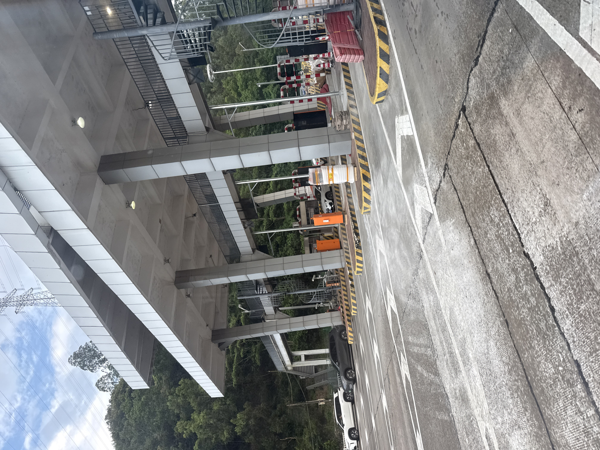 The image shows a toll plaza on a road in Huizhou, China. The scene is captured from a slightly elevated perspective, looking down at the toll booths and the road surface. The toll plaza structure is modern, with a large, elevated roof supported by multiple concrete columns. Various components of the toll collection system are visible, including barriers, signage, and what appear to be electronic payment systems. Yellow and black striped safety markings are present on the road and around the toll booths, indicating a hazardous area.
There are a few vehicles visible at the toll booths, including a dark-colored car and a white car. The road surface is asphalt, showing signs of wear and tear with cracks and patches. The surrounding environment is lush and green, with dense trees visible behind the toll plaza, suggesting a natural or semi-rural setting. Power lines and a transmission tower are also visible in the distance, indicating the presence of electrical infrastructure.
The lighting suggests it might be daytime, possibly overcast or in the late afternoon, as there are no strong shadows. No people are clearly discernible in the image, though there might be some figures in the background within the toll booth areas. No legible text is visible in the image. The overall impression is of a functional infrastructure setup within a natural landscape.