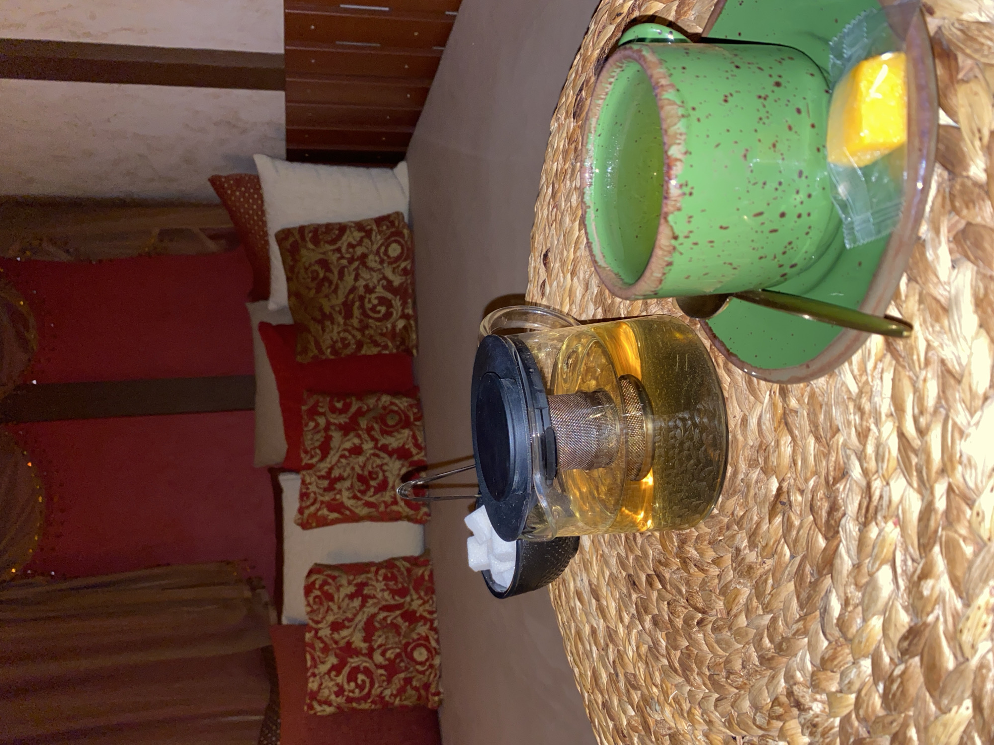 The image depicts a cozy, indoor scene, likely a living room or lounge area, with a focus on a tea set and decorative pillows. The overall ambiance suggests relaxation and comfort.
The primary objects are a green ceramic teacup with speckled detailing and a matching saucer, a glass teapot filled with amber-colored liquid, and a woven placemat. The teacup holds a green beverage, and a single cube of something yellow, possibly lemon or candy, is placed on the saucer, wrapped in clear plastic. A spoon rests on the saucer as well. The teapot appears to be made of glass with a black lid and a metal infuser. Next to the teapot are two small, white sugar cubes. The placemat is intricately woven from a natural material, possibly straw or rush, with a warm, golden-brown hue.
In the background, several decorative pillows are visible. Some are solid red, while others feature an ornate pattern of gold swirling motifs on a red background. White pillows are also present. The arrangement of the pillows suggests they are on a sofa or a seating area. Behind the seating area, there is a wall with horizontal wooden elements and a textured, light-colored section.
The lighting in the image is soft, contributing to the calm and inviting atmosphere. There is no visible text in the image. The scene implies a moment of quiet enjoyment, perhaps a tea break or a relaxing afternoon. The setting and the presence of the tea set suggest a comfortable and well-appointed indoor space.