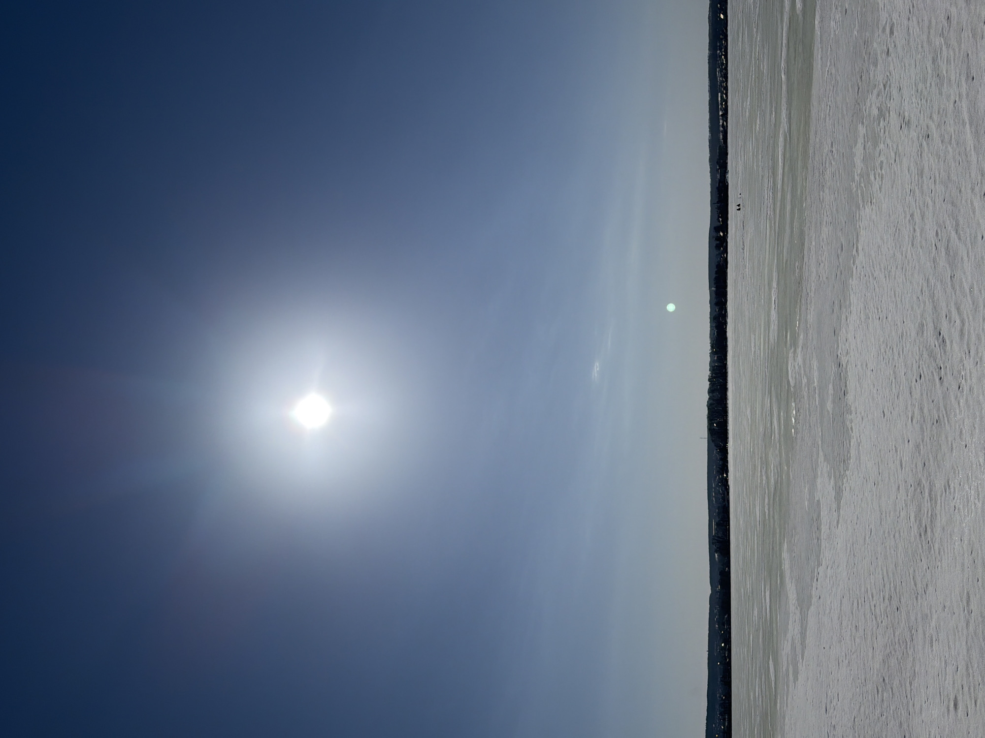 The image shows a winter scene, likely in Votkinsk, Russia. The horizon is horizontal in the image. The left side of the image is a blue sky with a bright sun. The right side shows a snow-covered expanse, probably a frozen body of water like a lake or river. Along the horizon line, there appears to be a line of trees or possibly buildings, which could be the cityscape of Votkinsk in the distance. Two tiny figures on the snowy ice are visible, they could be people walking or ice fishing. The surface of the frozen body of water has patterns that suggest ice and snow texture.