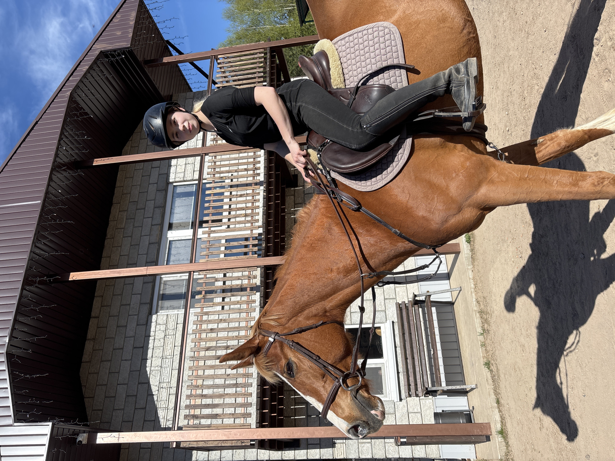A chestnut-colored horse stands with a rider on its back in Русский Вожой, Russia. The rider, wearing a black riding helmet, black shirt, and black riding pants, is seated in a brown leather saddle with a quilted pad. The horse has a brown leather bridle. The location appears to be outdoors, possibly a riding area near a two-story building with brown siding and grey brick walls. The photo is taken at a low angle, possibly from the ground or slightly below eye level. Sunlight is present, casting a shadow of the horse and rider on the ground.