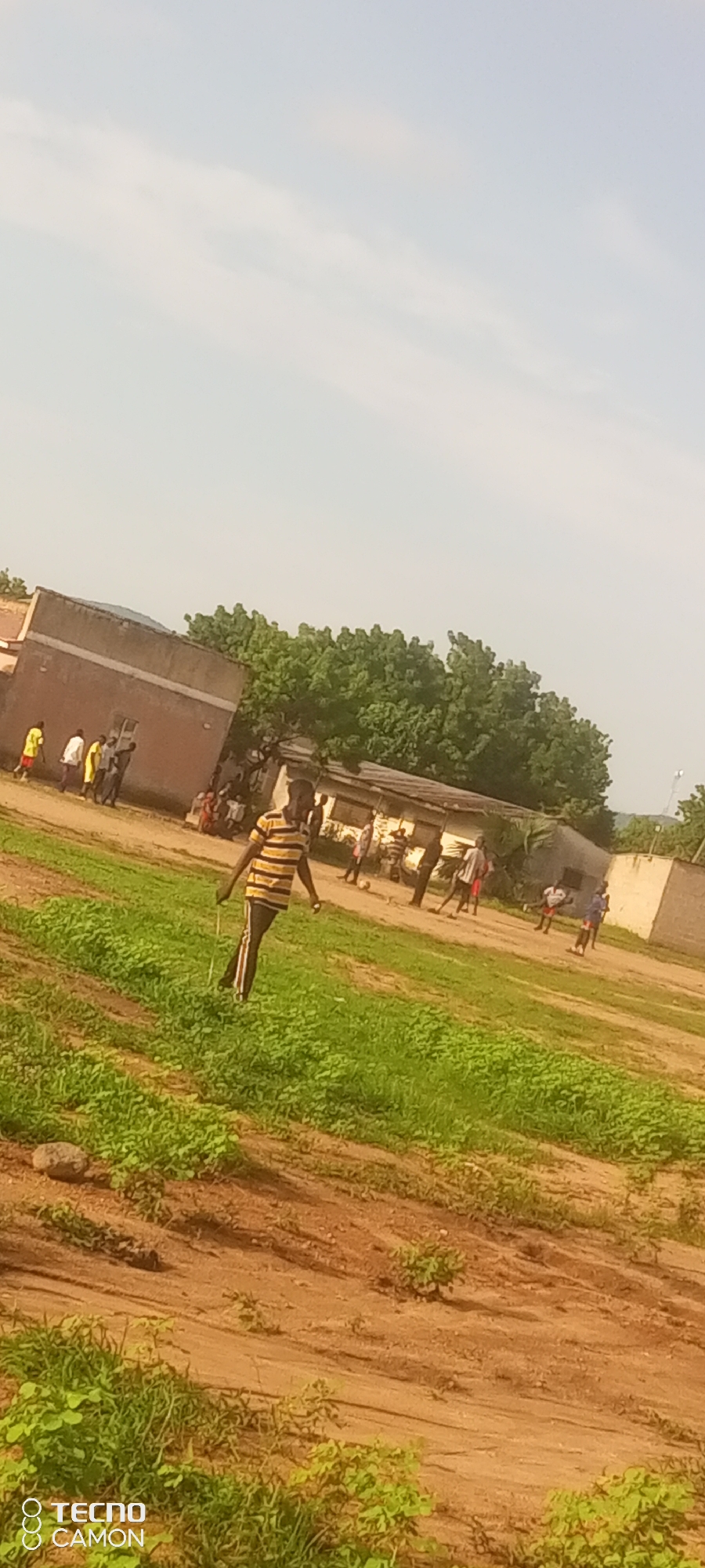The image depicts an outdoor scene with a group of people in Jalingo, Nigeria. In the foreground, a young male wearing a horizontally striped yellow and black shirt and dark pants with red stripes walks diagonally across a grassy field. He is holding a thin, light-colored stick horizontally in his right hand. The field has patches of bare earth and sparse green vegetation.
In the background, several buildings with light-colored walls and some with corrugated metal roofs are visible. A line of trees with dense green foliage forms a backdrop behind the buildings. A group of people, some appearing to be children or young adults, are gathered near the buildings. Several individuals are standing or walking, and some appear to be engaged in an activity, possibly a game, near a soccer ball. The sky is a pale blue with faint white clouds. A stone is visible in the lower left portion of the grassy area. The image also includes a watermark in the lower left corner with "TECNO CAMON" text and logos.