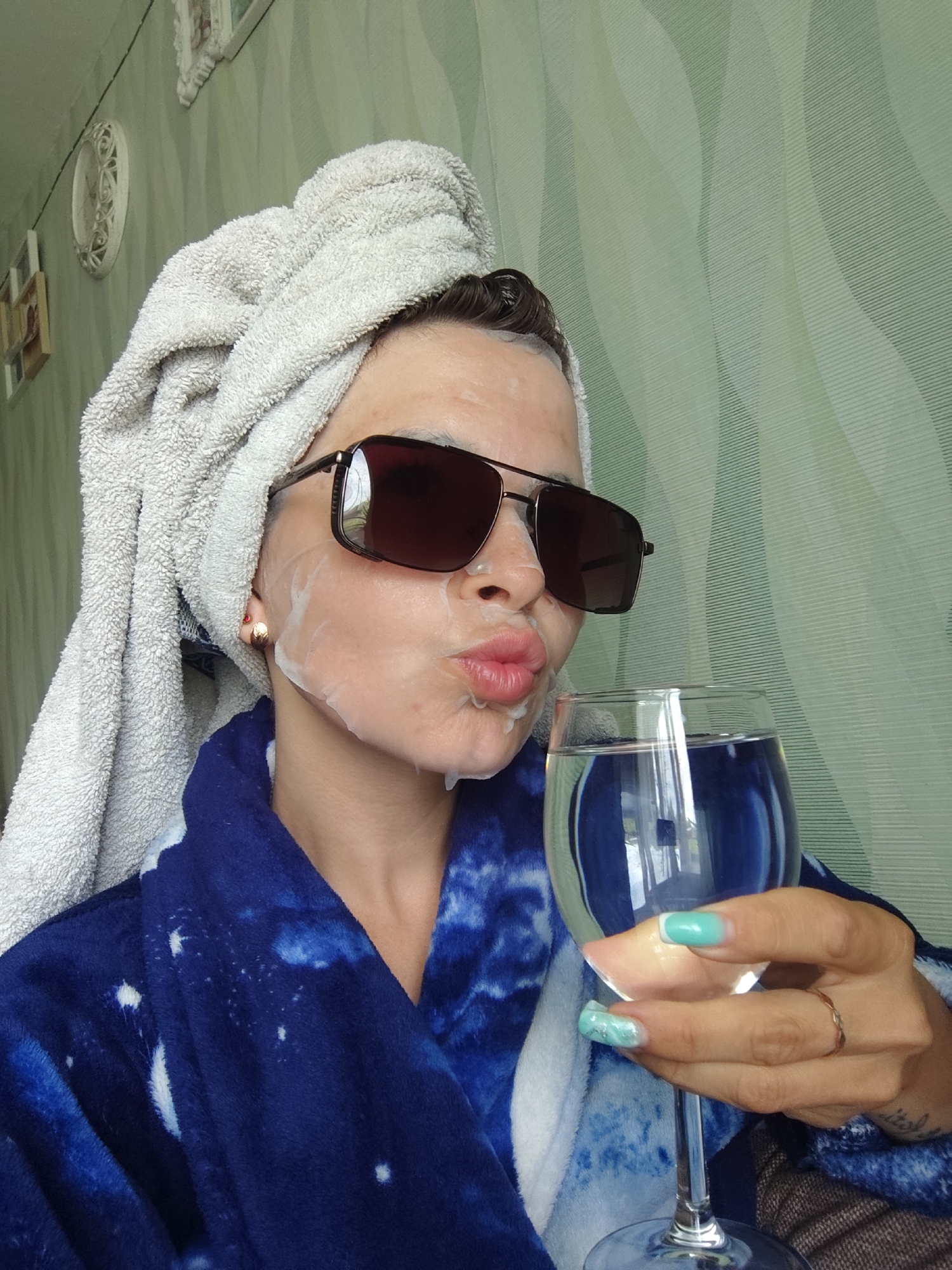 A person in a blue and white patterned robe and a white towel wrapped around their head is holding a glass of water, posing for a selfie. They are wearing sunglasses and have a white substance on their face, possibly a face mask. The person's nails are painted in a marbled turquoise and white pattern, and they wear a simple gold ring on their ring finger. The background is a light green wall with a wavy pattern. A picture frame and an oval clock are visible on the wall in the upper left corner. The location is Фоки, Russia.