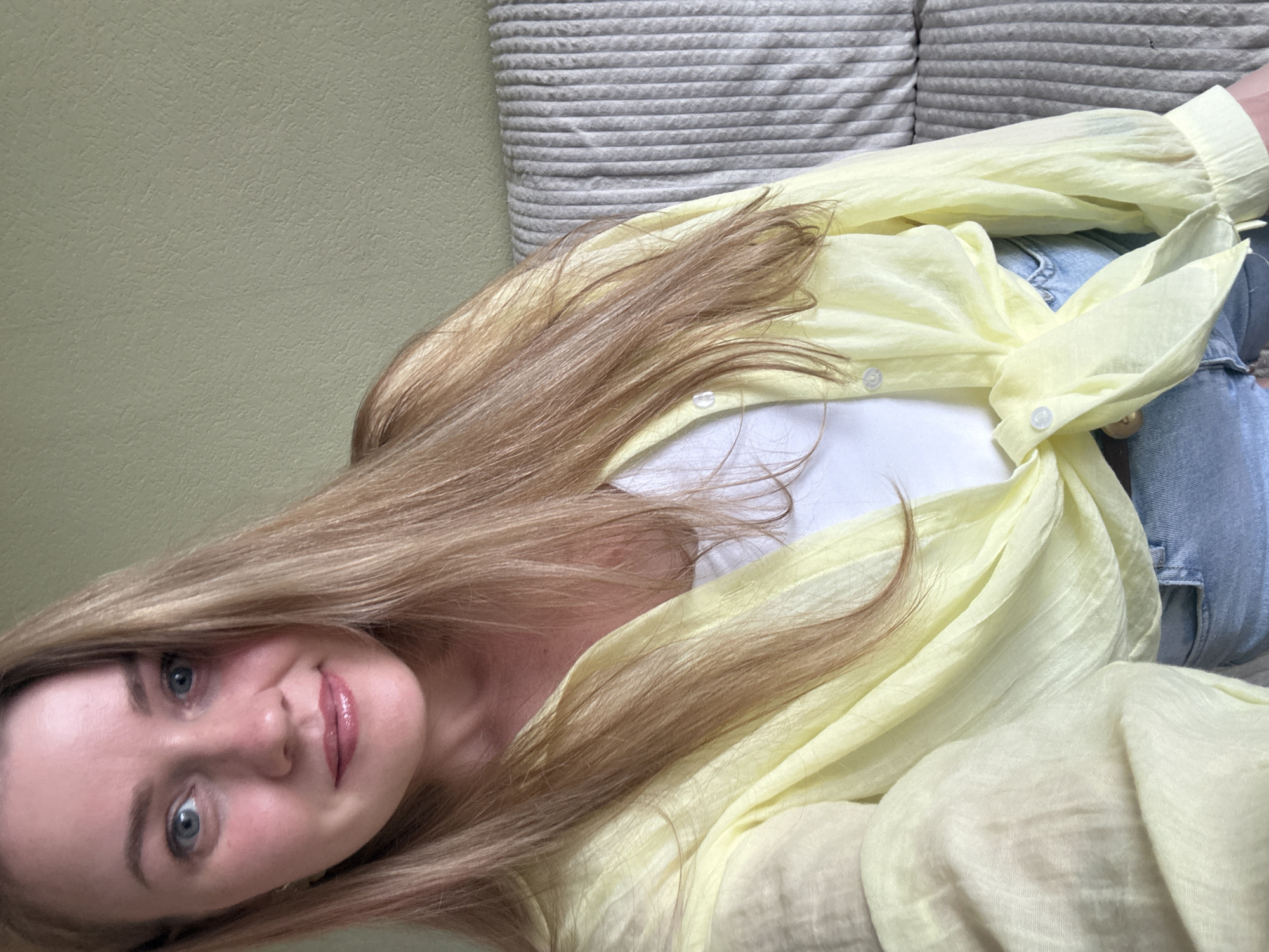 In a selfie taken indoors, a woman with long, blonde hair and blue eyes looks directly at the camera. She is wearing a light yellow, unbuttoned shirt tied at the waist over a white tank top, and light-wash denim shorts. Her makeup includes pink lipstick and mascara. She is seated, with her body angled slightly to the right, and the background shows a textured, light green wall and a portion of a ribbed, grey sofa. The image does not provide any visual cues to determine a specific location within Chaikovsky, Russia.