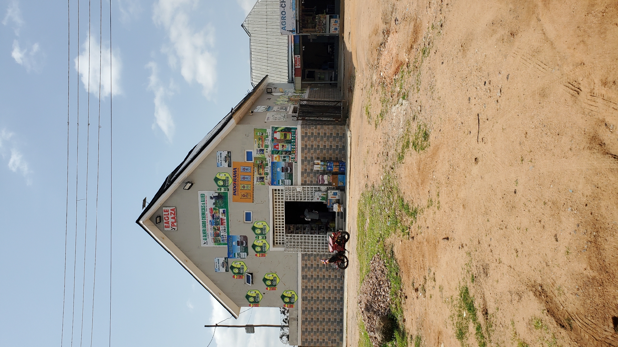 The image captures a frontal view of a commercial building in Jalingo, Nigeria, identified by the signage "EAGLE PLAZA". The building is a single-story structure with a prominent gabled roof, painted a light gray. It appears to be an agricultural supply store, as indicated by numerous advertisements for agrochemicals and related products displayed on its exterior walls. The front of the building features an open storefront with a doorway, revealing shelves stocked with various items inside. A man is partially visible standing within the store.
To the right of the main building, a smaller, connected structure with a corrugated metal roof extends outwards, also housing commercial activity with signage reading "AGRO-CH...". The area in front of the buildings is a dusty, unpaved ground, characteristic of many roads and commercial areas in Nigerian cities. A red motorcycle is parked in front of the main storefront, and stacks of products are visible near the entrance. The environment is illuminated by daylight, with a partly cloudy sky overhead. The overall scene depicts a typical commercial street in Jalingo, focusing on a business that likely serves the agricultural community.