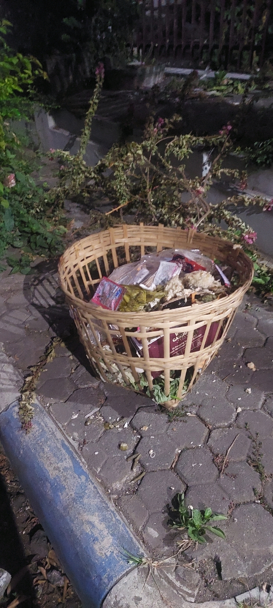 In Lumajang, Indonesia, a woven bamboo trash bin is positioned outdoors on a paved walkway. The bin is overflowing with a variety of refuse, including plastic packaging, paper items, and organic waste. The surrounding area is dimly lit and features lush green foliage and some pink flowers, suggesting a garden or park setting. The ground is paved with interlocking hexagonal stones, and a blue-painted curb is visible in the foreground. Scattered debris, such as leaves and small twigs, are present on the walkway. The overall impression is one of a somewhat neglected outdoor space, with the trash bin serving as a focal point.