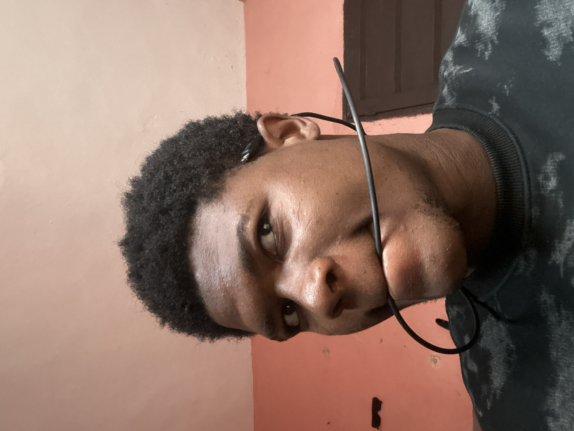 The image shows a close-up, head-and-shoulders view of a young Black man with short, dark, curly hair and dark skin. He is looking to his left with his mouth slightly open, biting down on a black cable, likely from earphones, which is draped around his neck and over his shoulder. The background is a plain, textured wall with a reddish-pink hue. The man is wearing a dark-colored sweatshirt with a light-colored, abstract pattern. The lighting is soft, creating some subtle shadows on his face. The scene appears to be indoors, possibly in a residential setting in Ihiagwa, Nigeria, suggested by the wall texture and the man's appearance.