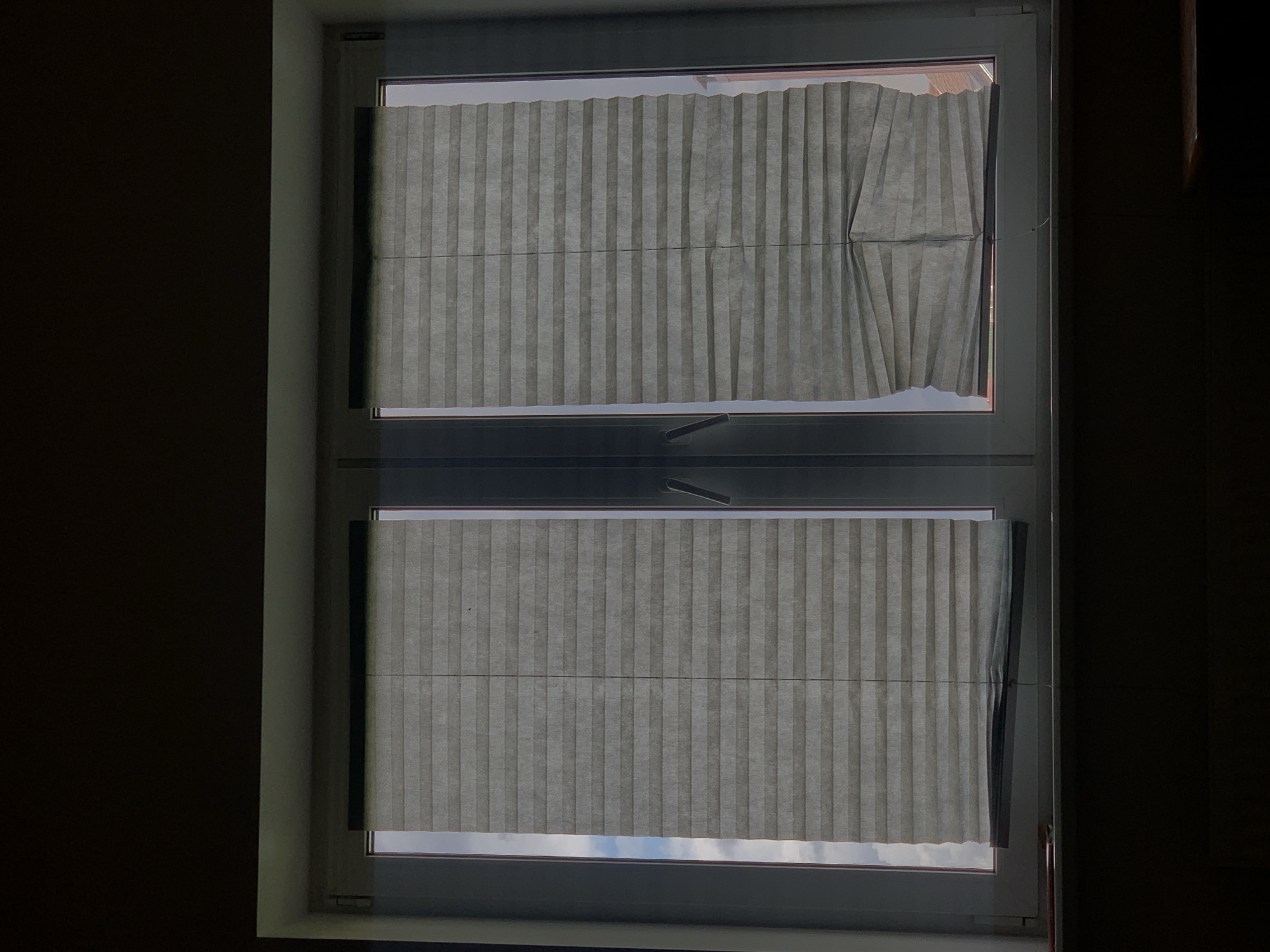 The image shows a window with two pleated blinds. The window is divided into two vertical panes. The left blind is fully extended, while the right blind is partially lowered and appears to be bunched up at the bottom. The blinds are light gray and translucent, allowing some light to filter through. Outside the window, the sky is visible with some clouds. The window frame is white. The overall scene is dimly lit, with the majority of the image in shadow. There are no people or animals visible in the image. The context of Minsk, Belarus, is not discernible from the visual information presented in the image itself.