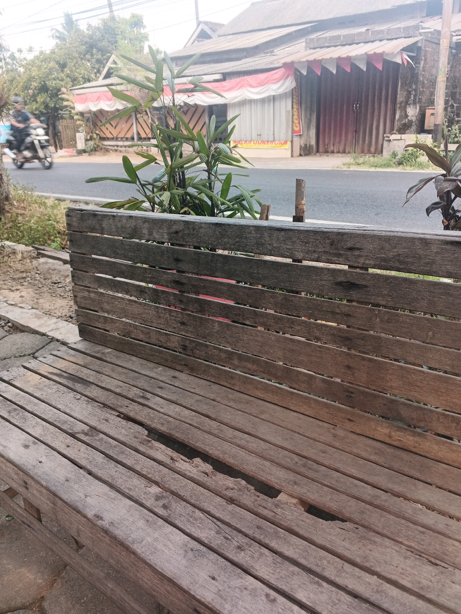 The image depicts a weathered wooden bench in the foreground, with its slats showing signs of wear and a broken section on the seat. Behind the bench, a road runs horizontally across the frame. On the other side of the road, a building with a corrugated metal roof and a red and white awning is visible. The building appears to be a storefront, with metal shutters closed. A motorcycle carrying a person is visible in the distance on the road, moving from left to right. Green foliage, including a small plant in a planter box on the bench, and trees are present in the scene. The overall environment suggests a casual outdoor setting, possibly in a semi-urban area of Ungaran, Indonesia, given the architectural style of the building and the presence of a motorcycle.