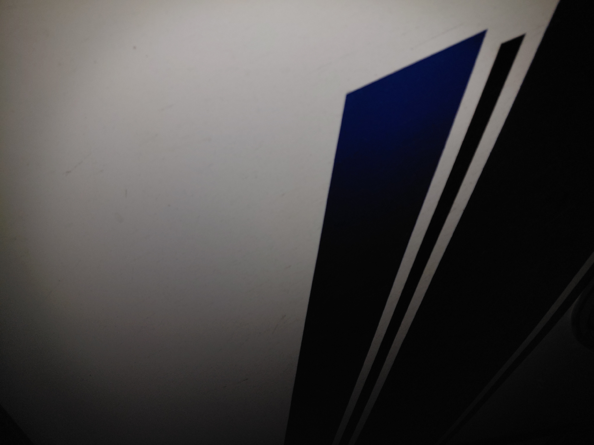 The media file displays an abstract, close-up view of a geometric design featuring distinct color blocks and parallel lines. The composition is dominated by a large, light gray or off-white area occupying the upper left quadrant. This surface appears to have subtle textures or minor imperfections.
Diagonally cutting across the image, a sharp edge separates the light gray area from a deep, rich blue triangular or trapezoidal shape. This blue segment then meets a broad black stripe, which is itself bordered by a pattern of two thin, parallel white lines set against a dark black background. The lines and color blocks are oriented diagonally, sloping downwards from left to right.
The lighting is uneven, creating a gradual vignette effect where the top-left portion is brighter, and the image progressively darkens towards the lower right. There are no people or specific activities depicted. No text is visible. The image captures a static detail, possibly part of a stylized emblem, logo, or decorative element on a larger object, within the context of Santa Ana, Venezuela.
