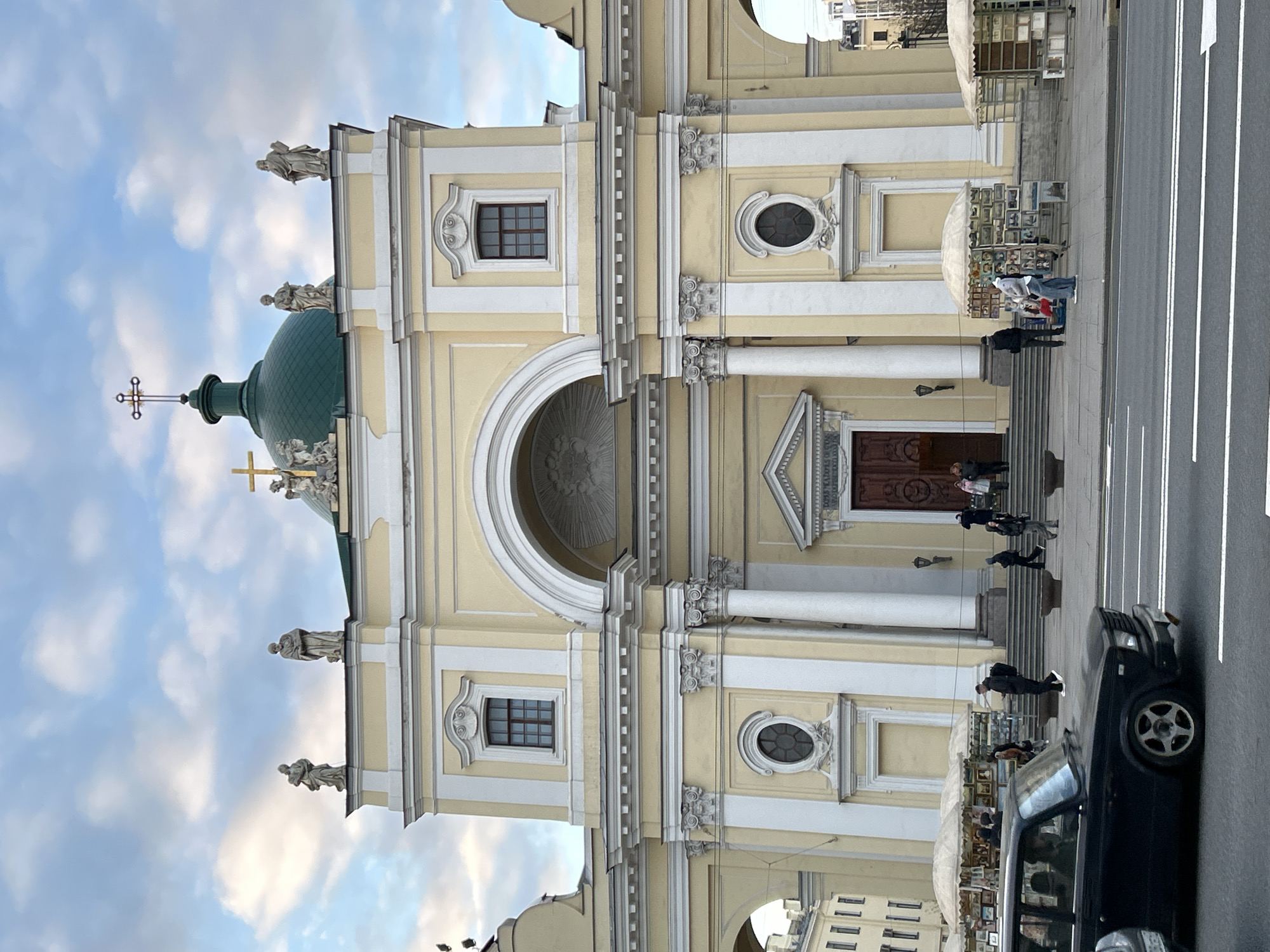 The image captures the facade of a large, ornate church in Saint Petersburg, Russia, under a partly cloudy daytime sky. The church building is predominantly light yellow with white architectural details, including pilasters, columns, cornices, and decorative elements. A prominent green dome, featuring a golden cross and topped with an ornate metal cross, rises from the roofline. Several statues of figures, likely saints, adorn the roofline and niches on the facade.
The church features a grand central arched entrance, with what appears to be a smaller, rectangular window above. To the right of the central arch is another entrance with large wooden double doors, above which an inscription reads "S. ANTONII DE PADVA," indicating its dedication to Saint Anthony of Padua. Windows on the facade are a mix of rectangular and oval shapes, some with ornate surrounds.
In front of the church, a paved street runs parallel to the building, with a wide sidewalk separating it from the church steps. Several people are visible on the steps leading to the church and on the sidewalk. An active outdoor market is set up on the sidewalk, with multiple white-canopied stalls displaying numerous framed pictures or artworks for sale. People are seen browsing these stalls. A dark-colored car is partially visible on the street in the foreground, suggesting urban activity.
The lighting is bright, indicating a clear day, possibly late morning or early afternoon. The overall scene depicts a bustling urban environment where religious architecture meets daily city life and commerce.