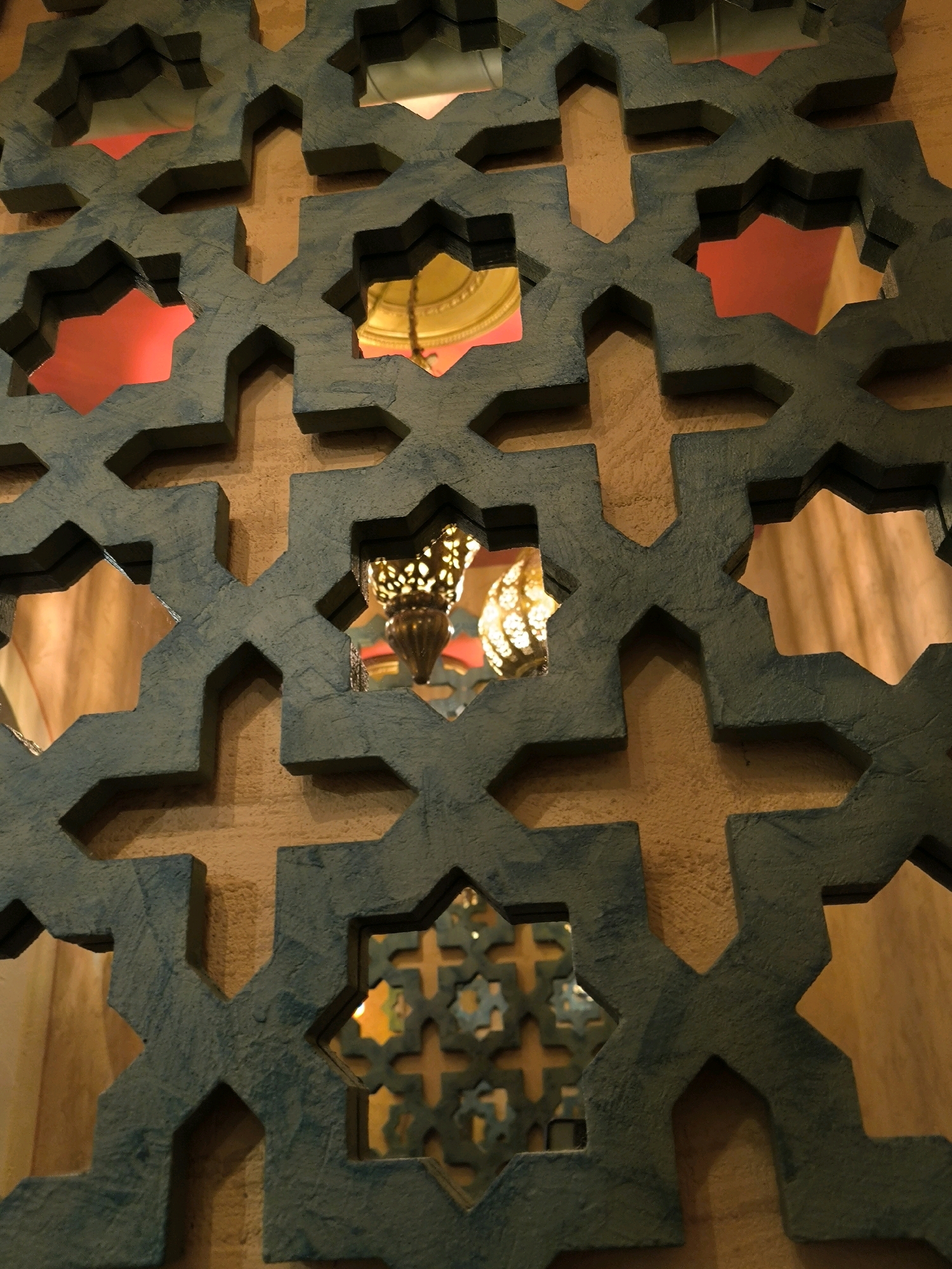The image shows a close-up view of a decorative screen with an intricate geometric pattern. The screen is made of a dark teal material and is mounted against a textured, light brown wall. The pattern consists of interlocking star shapes and crosses, creating openings through which glimpses of the interior can be seen.
Through the openings, a warm, golden light is visible, suggesting interior lighting or possibly a light source behind the screen. In some of the openings, decorative lamps are visible. One lamp, in particular, appears to be a hanging pendant light with a perforated metal shade, emitting a patterned light. Other openings reveal solid red and plain golden surfaces, possibly painted walls or fabric. The overall aesthetic suggests an indoor environment, possibly a public space or a room with Middle Eastern or Islamic architectural influences.
There are no people or animals visible in the image. The focus is entirely on the architectural detail of the screen and the interplay of light and shadow through its openings. The image is taken from a slightly downward angle, looking through the screen.
This image was taken in Novosibirsk, Russia. Specifically, it is a detail from the interior of a restaurant called "Ufigr" (Уфигр) located at 23 Komsomolsky Prospekt, Novosibirsk. The restaurant is known for its interior design inspired by Oriental motifs, which includes decorative screens like the one pictured.