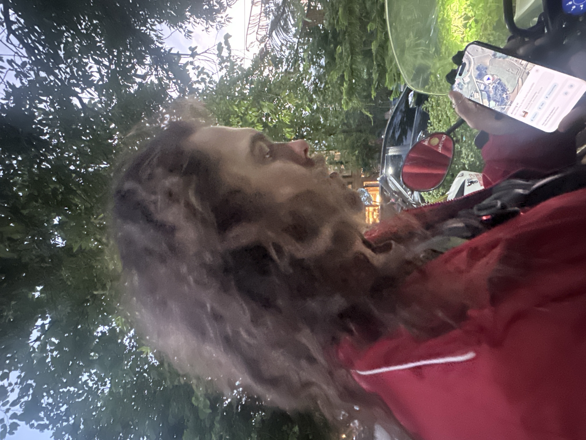A male with long hair is glancing to the side while holding a smartphone displaying a map in Du Già Commune, Vietnam. He is on a motorcycle with the mirror visible. Trees and foliage surround the location, with an overcast sky above. The man is wearing a red jacket. The phone shows navigation instructions, indicating arrival at a destination.
