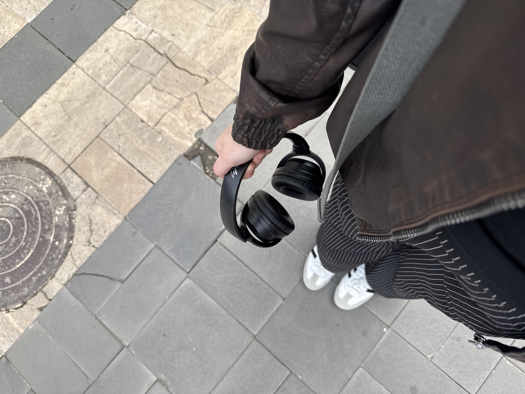 A person wearing a brown leather jacket, striped pants, and white sneakers stands on a paved surface in Yerevan, Armenia. They are holding a pair of black over-ear headphones in their right hand. The pavement is composed of grey and tan rectangular blocks. A circular manhole cover is visible in the upper left corner of the frame.