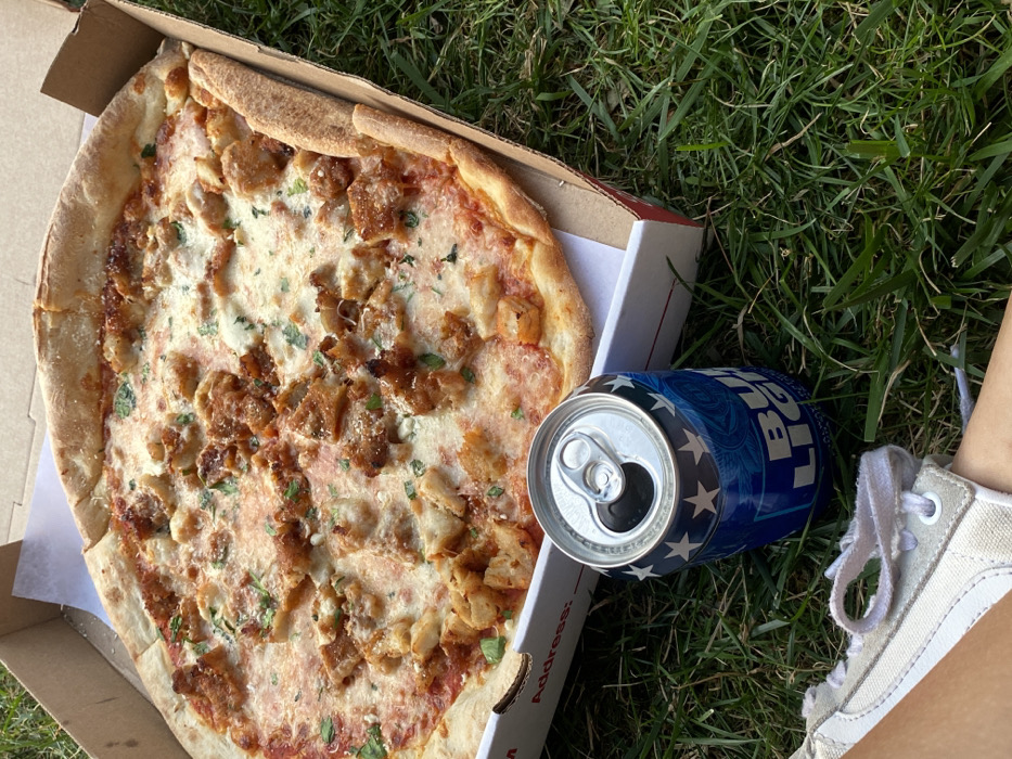 The image depicts an outdoor scene in Ridgefield, United States, focusing on food and drink placed on green grass. A large, round pizza occupies a significant portion of the frame, resting inside an open, standard brown cardboard delivery box lined with white paper. The pizza features a golden crust, red tomato sauce, melted white cheese, and numerous irregularly shaped pieces of cooked chicken as a topping, some appearing breaded or fried. Green chopped herbs are sprinkled over the pizza. To the left of the pizza box, an open, blue and white Bud Light beer can, featuring stars and brand text, stands upright on the grass. In the lower left quadrant of the image, the lower leg and foot of a person are visible, wearing a light-colored, lace-up sneaker, indicating a human presence sitting on the grass. The grass is green and appears well-maintained, suggesting a lawn or park setting under natural daylight.