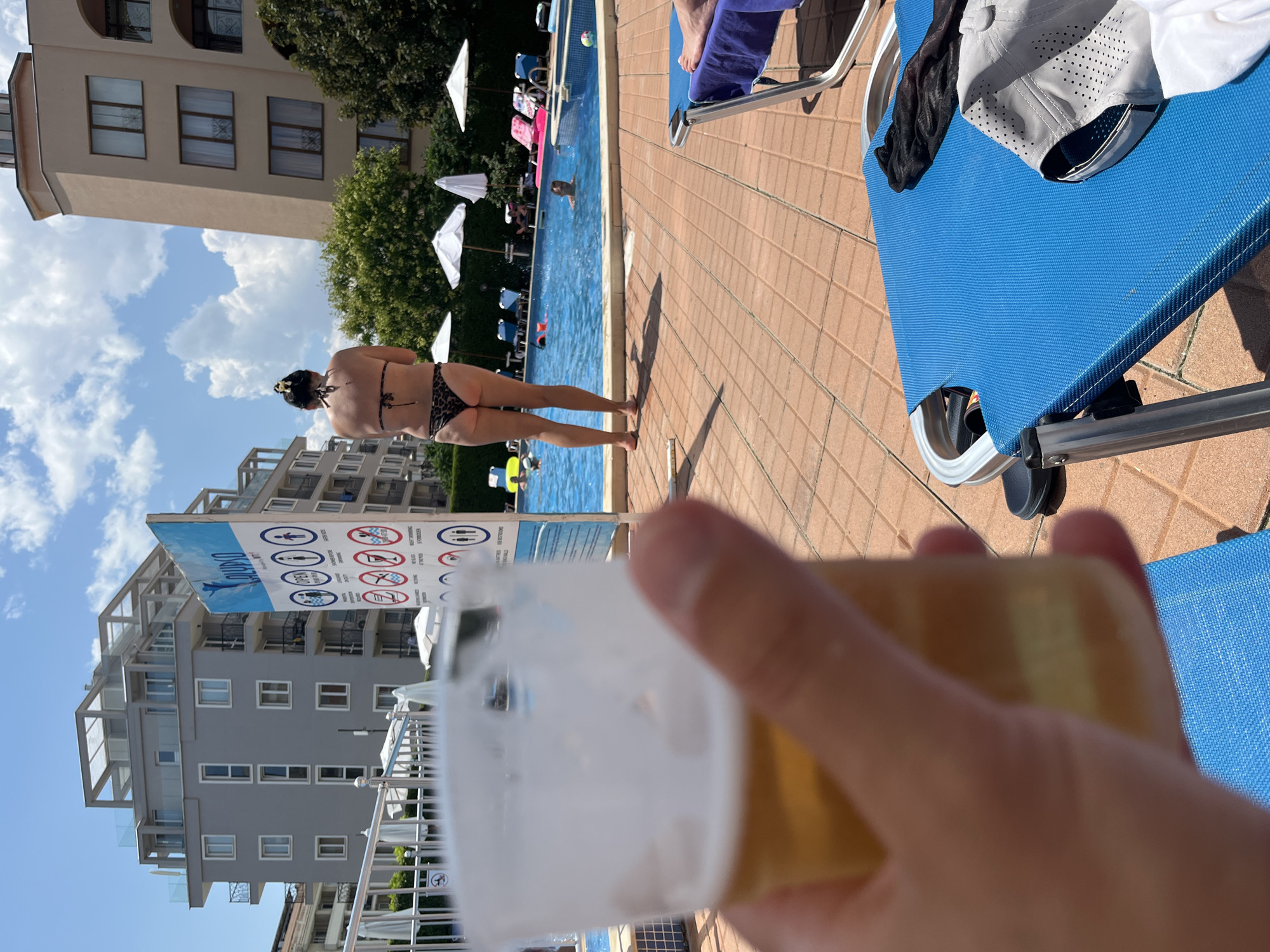A person's hand holds a blurred plastic cup of beer in the foreground. In the midground, a person wearing a leopard print bikini stands facing away from the camera, looking towards a swimming pool. Beyond the pool, buildings are visible. A sign with rules in pictograms is next to the pool. The scene appears to be at a hotel or resort in Nesebar, Bulgaria, indicated by the sign's text in Cyrillic, which reads "CALYPSO". The area is paved with terracotta tiles, and lounge chairs are set up around the pool. The sky is blue with scattered clouds.