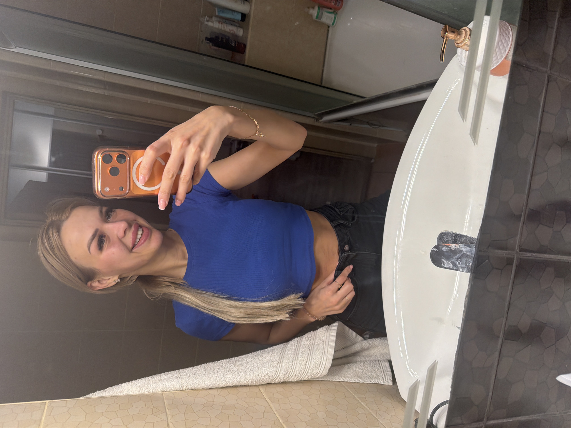 A young woman with blonde hair pulled back in a ponytail, wearing a blue crop top and black jeans, takes a selfie in a bathroom mirror. She is smiling and holding her phone. The bathroom appears to be in an apartment, with tiles on the walls and a sink visible in the foreground. Various toiletries are on shelves in the background. The overall scene suggests a casual moment captured at home.