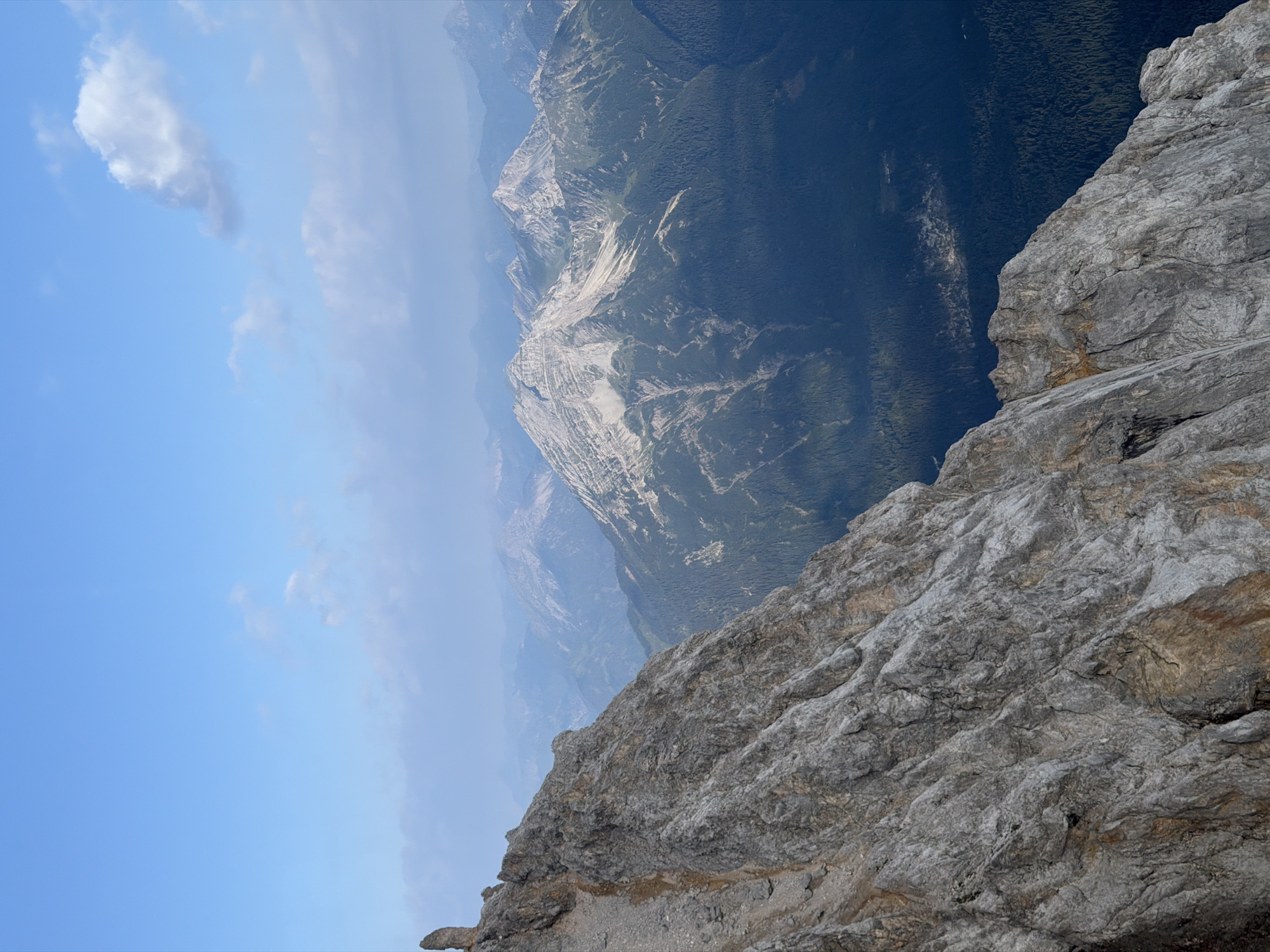 This image captures a panoramic view of the Zugspitze, Germany's highest peak, as seen from a rocky mountain ridge. The foreground is dominated by a steep, grey, rocky slope with visible texture and some patches of brown earth. In the mid-ground, a series of majestic, rugged mountains rise, some with bare rock faces and others covered in dark green coniferous forests. The distant mountains appear hazy and are layered in shades of blue and grey, indicating depth and atmospheric perspective. The sky above is a clear, bright blue with a few scattered, wispy white clouds. The scene suggests a high-altitude, natural environment within the Bavarian Alps, likely near Grainau, Germany, given the prominence of the Zugspitze. No people or animals are visible in the frame.