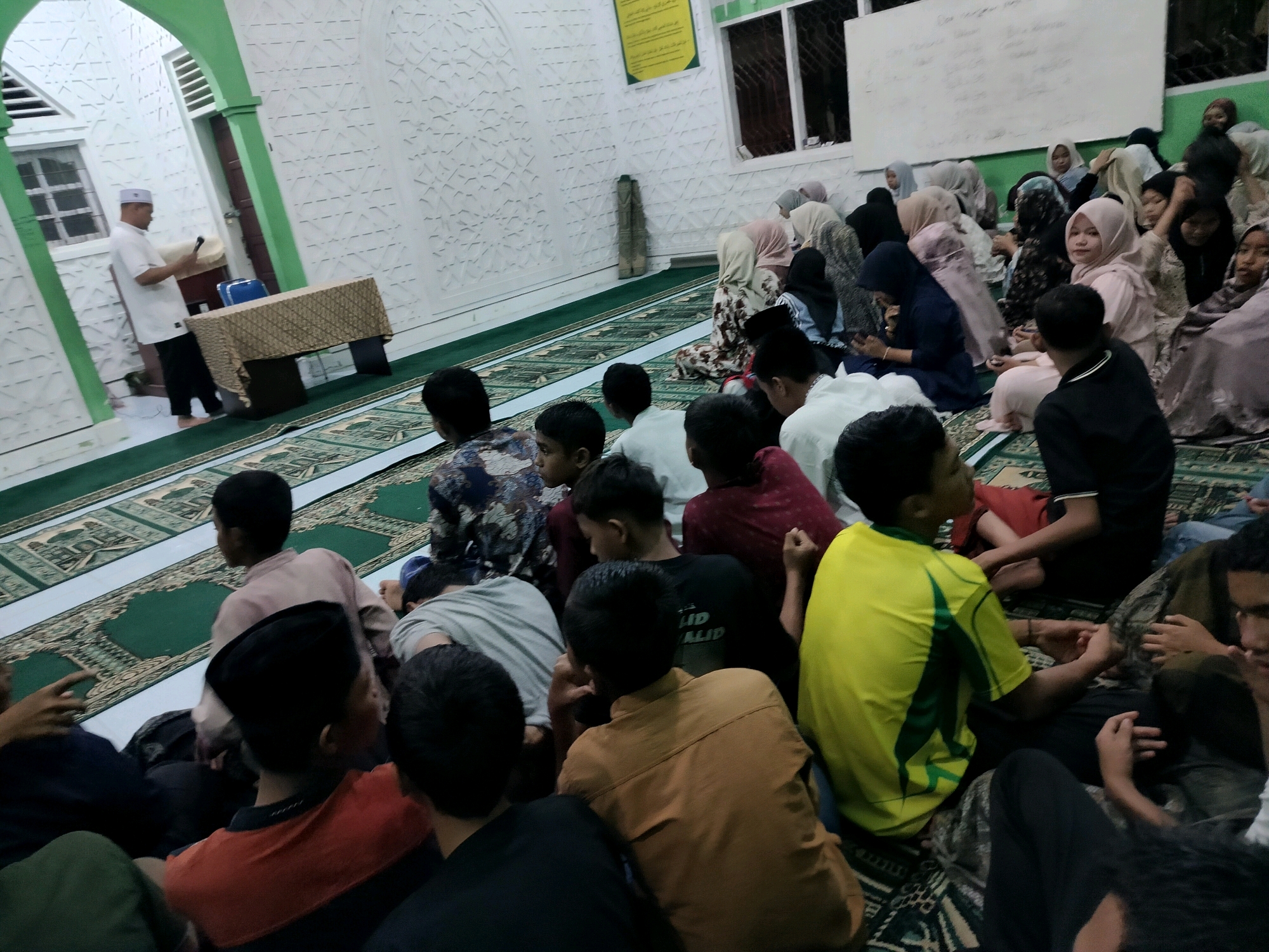 The image depicts an indoor gathering of people, likely at a mosque or community hall, in Purwodadi, Indonesia. In the foreground, a group of young males are seated on prayer mats, facing away from the camera. One individual in a yellow shirt with green accents is prominent. To the left, a man in a white shirt and cap stands near a table covered with a patterned cloth, holding a device that could be a microphone or phone. In the background, a larger group of people, predominantly women and some children, are seated on prayer mats. They are dressed in modest attire, many wearing hijabs. The walls of the space are decorated with intricate white geometric patterns. The environment suggests a religious or educational event, possibly a lecture or prayer session, given the seating arrangement and the presence of prayer mats. The lighting is somewhat dim, with artificial light sources illuminating the scene.