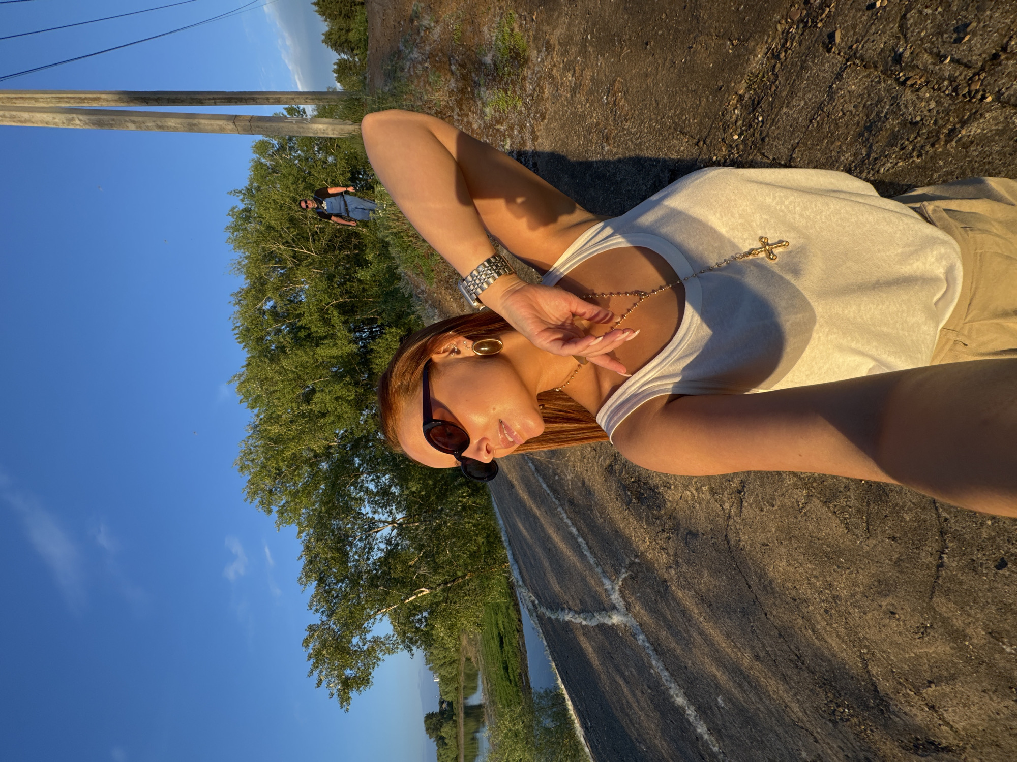 An outdoor, medium-shot, selfie-style image depicts a woman standing on a rocky embankment, illuminated by the golden light of a setting sun. She is wearing a white tank top, beige pants, large oval sunglasses, and a silver watch. A gold necklace with a cross pendant is visible. Her right arm is bent and resting on her head, and her left arm is outstretched holding the camera. In the background, a river and green trees are visible, with a man in denim overalls walking in the distance. Two large utility poles stand to the right of the frame, along with power lines. The location is identified as Chaikovsky, Russia, near the Kama River.