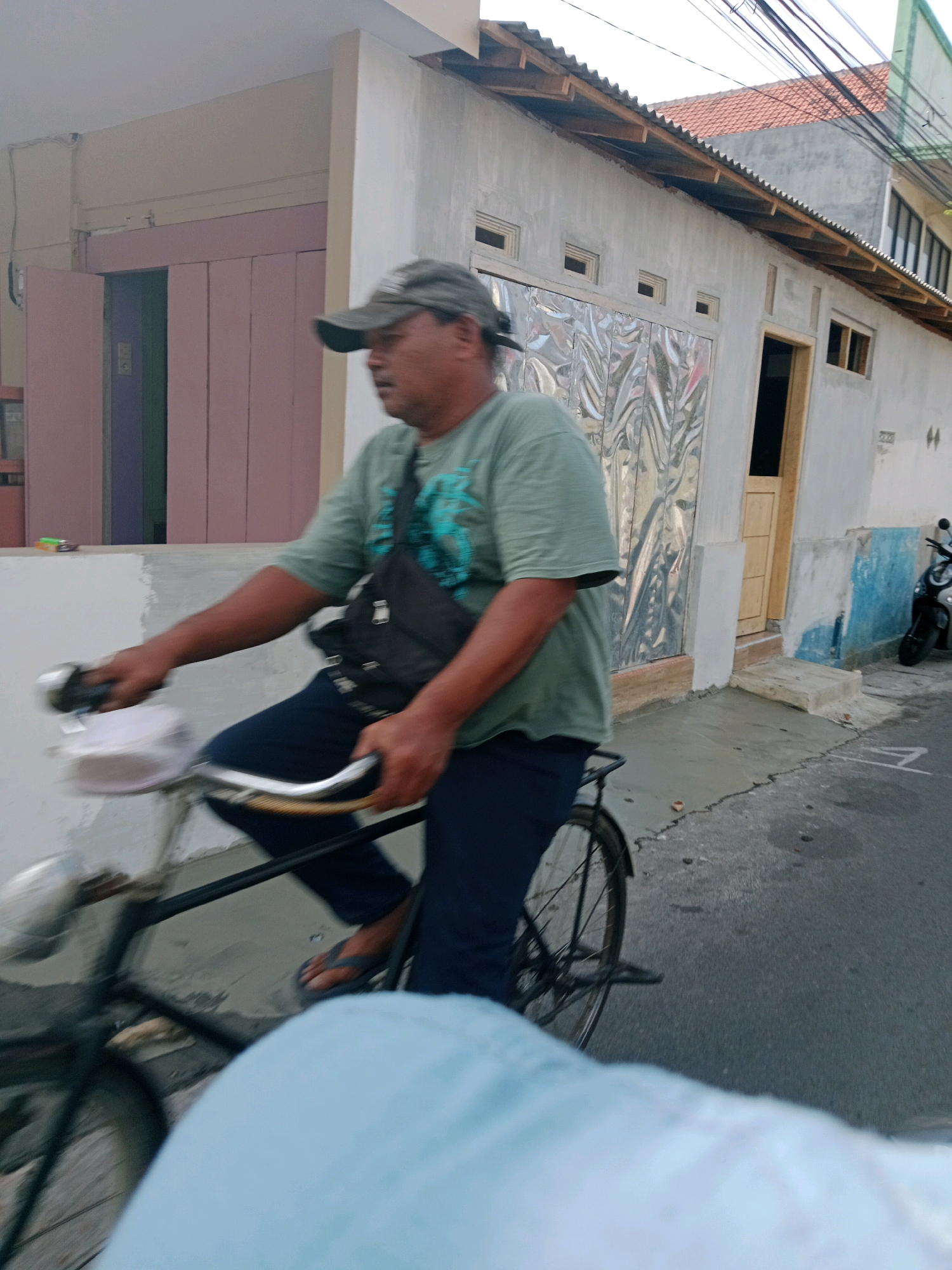 A man wearing a green t-shirt, dark blue pants, and a grey baseball cap is riding a bicycle on an asphalt road. He has a black crossbody bag slung over his shoulder. The bicycle has a white basket on the front. In the background, there is a building with a white facade, a brown wooden door, and silver-colored corrugated metal panels. A motorcycle is parked on the right side of the road. The scene appears to be in Probolinggo, Indonesia, as suggested by the architectural style and the presence of a motorcycle commonly used in the region. The lighting suggests it is daytime, possibly late afternoon. The road is wet in some areas.