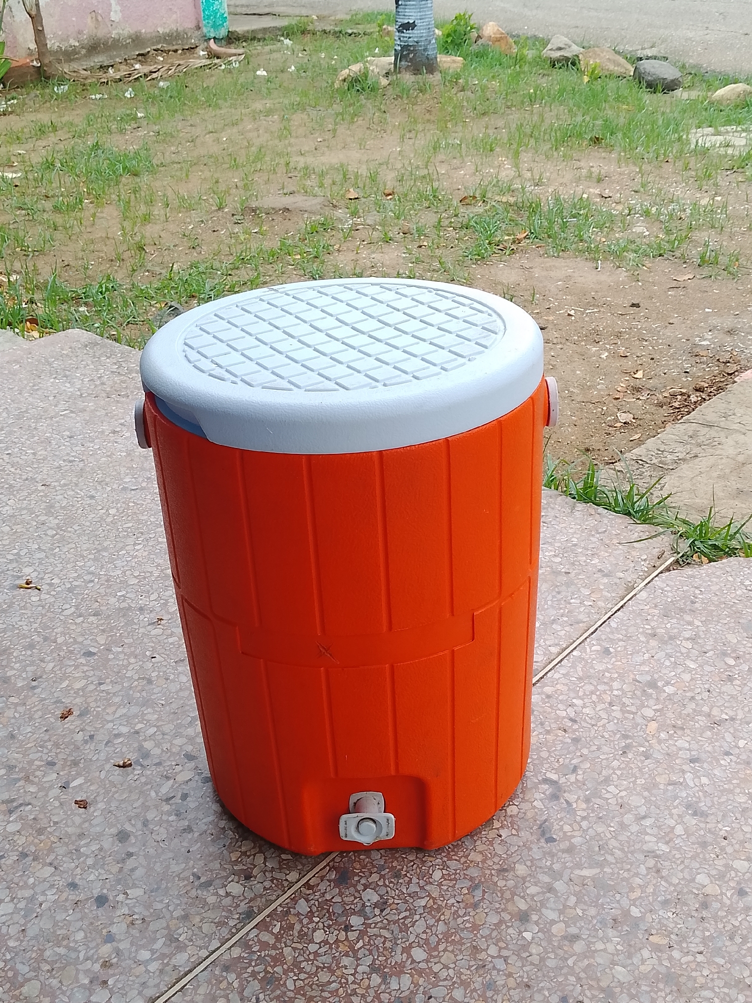 An orange water cooler with a white lid and a spigot is positioned on a speckled, grey, concrete surface outdoors. The cooler has two grey handles, one on each side. The background shows a grassy area with some scattered dirt, rocks, and a blurry, light-colored building. The scene appears to be in a residential or public outdoor space in Altagracia, Venezuela. There are no people visible in the image.