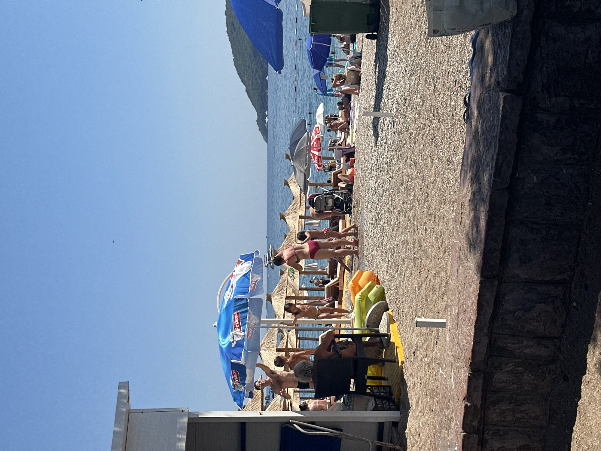 The image depicts a crowded beach scene in Budva, Montenegro, under a clear blue sky. The foreground is dominated by a textured wall with a white pole emerging from the gravelly ground behind it. The beach itself is filled with numerous people, many of whom are sunbathing, swimming, or relaxing under umbrellas.
In the middle ground, several beach umbrellas provide shade, with one large blue umbrella prominently featuring a logo. People are seen standing and sitting under these umbrellas. A man in sunglasses is standing and gesturing, while a woman in a bikini stands near him. Children are also present, with one girl standing near the blue umbrella. Further back, more umbrellas and beachgoers are visible, along with lounge chairs and a stroller.
The background shows the calm, blue sea, with a few boats in the distance and a lush green hill on the horizon. The overall impression is a lively and popular summer beach resort.