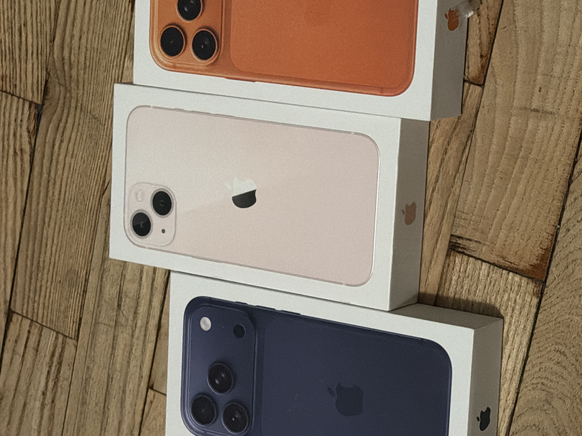 The image shows a stack of three iPhone boxes on a wooden surface. The top box features an orange iPhone 11 Pro/Max, the middle box features a white/silver iPhone 11, and the bottom box features a blue iPhone 12. The background appears to be a wooden floor. The location in the city of Varna, Bulgaria is not evident from the image.