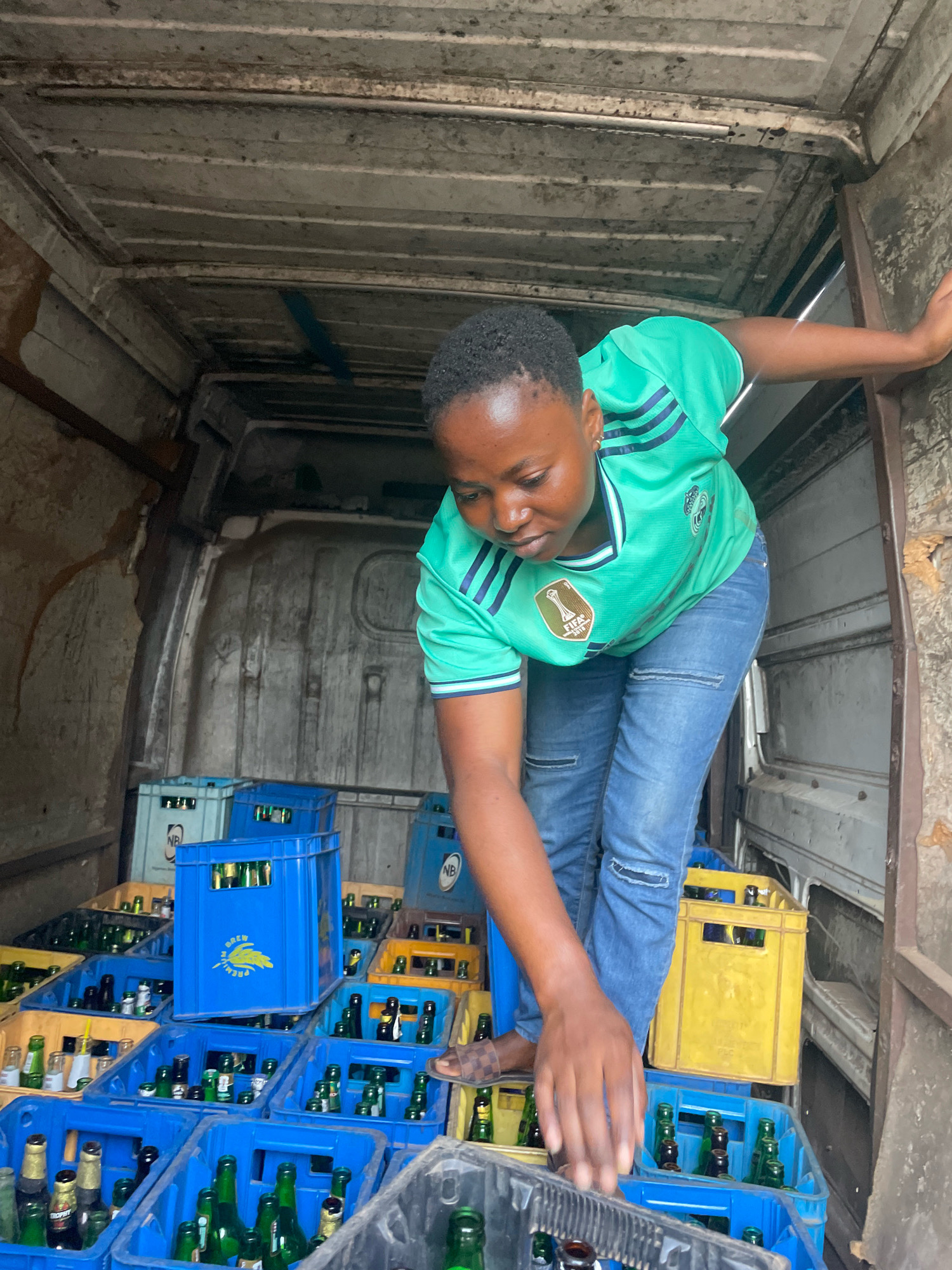 A woman, likely in her late twenties or early thirties, wearing a bright green soccer jersey and ripped blue jeans, is seen bending down inside the back of a van. She is reaching into a blue plastic crate filled with green glass bottles, presumably beverages. The van's interior is packed with crates of bottles, some blue and some yellow, stacked high. The environment appears to be a service area or loading dock, potentially in Jos, Nigeria, suggested by the context of this image set. The van's interior walls and ceiling show signs of wear and tear, with grime and discoloration. The woman's posture and focus indicate she is actively engaged in loading or unloading the bottles.