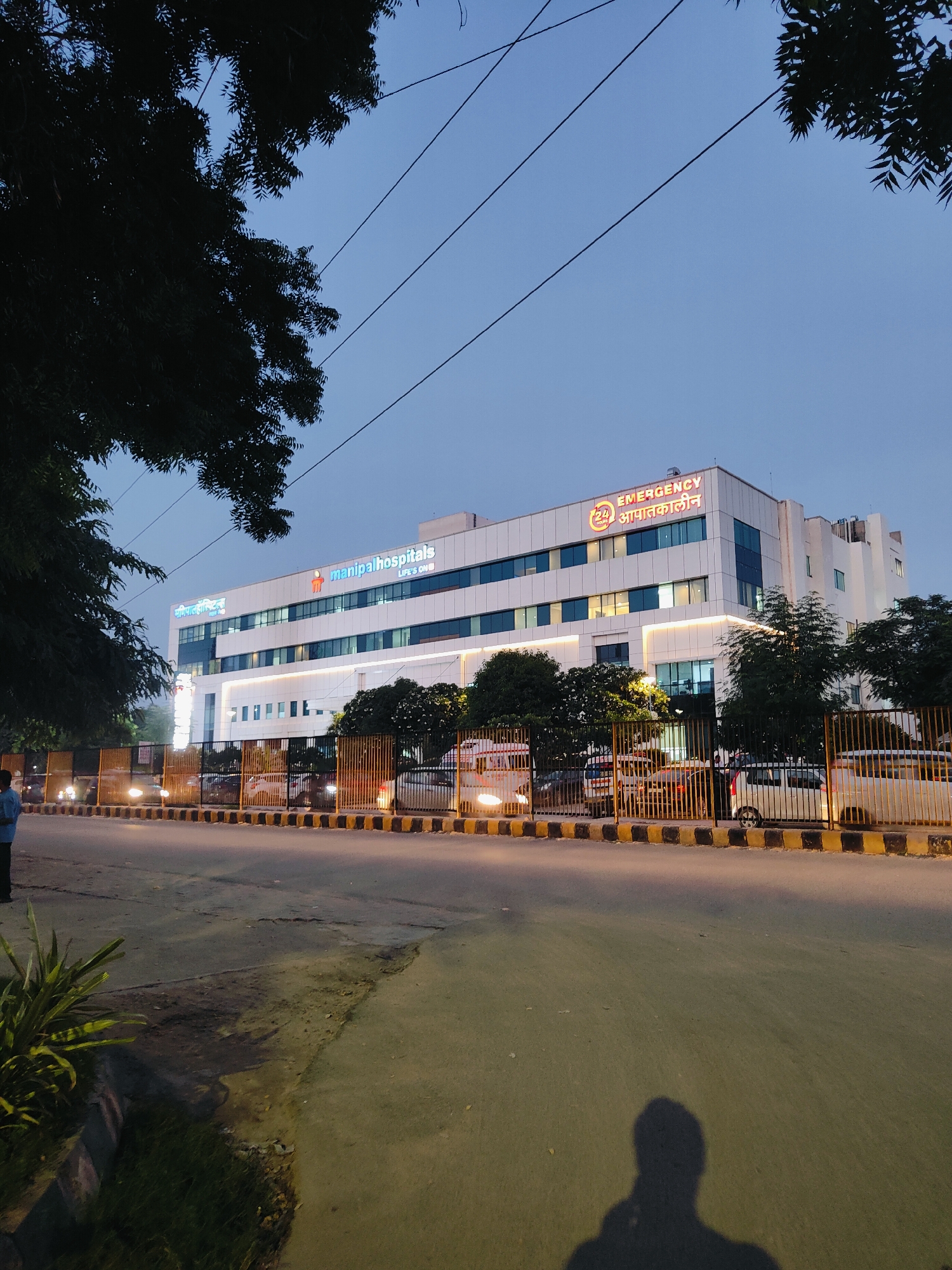 A wide shot captures the exterior of Manipal Hospitals in Gurgaon, India, at dusk. The multi-story building is illuminated, with a prominent "manipalhospitals" logo in blue and orange on its facade. Above, "24 EMERGENCY" is displayed in large, red lettering, accompanied by Hindi text. The building's modern architecture features rows of windows and white siding.
In the foreground, a street separates the viewer from the hospital. Cars are visible on the road, with their headlights on. A black and yellow striped barrier runs along the edge of the hospital property. Trees with dark foliage frame the left and right sides of the image. The sky is a deep blue, indicating the transition from day to night. A faint shadow of a person, likely the photographer, is cast on the road in the lower right corner. A single person, wearing a light blue shirt, stands on the sidewalk to the left. The overall scene depicts a well-lit hospital in an urban setting at twilight.