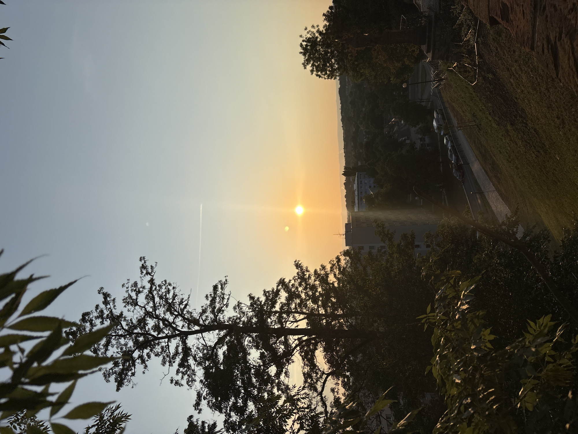 A high-angle shot captures a sunset over a cityscape. The sun, a bright orb, is positioned in the upper right quadrant of the frame, casting a warm, golden light across the sky. A faint contrail is visible in the sky above the sun. In the foreground, silhouetted trees and foliage frame the scene. Below, a street with parked cars winds its way through a dense arrangement of trees and buildings. The architecture appears to be a mix of modern and older structures. A grassy slope leads down from the viewer's vantage point towards the street and the distant horizon, which is marked by rolling hills under the fading light. The overall impression is one of a peaceful evening in an urban environment.