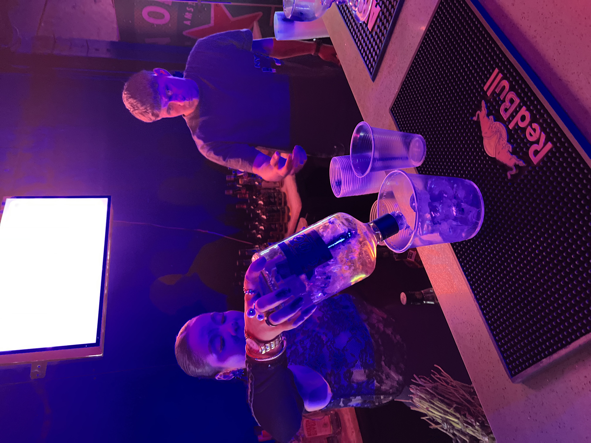 The image depicts a bar scene, possibly in Arica, Chile. A man stands behind the bar, wearing a long-sleeved shirt. In front of him are plastic cups and a bottle of alcohol which he is pouring into a cup filled with ice. Another person is visible in the lower left corner, wearing a dark top with lace. There is a Red Bull-branded mat on the counter, and a television screen is visible. A row of bottles are visible behind the first man.