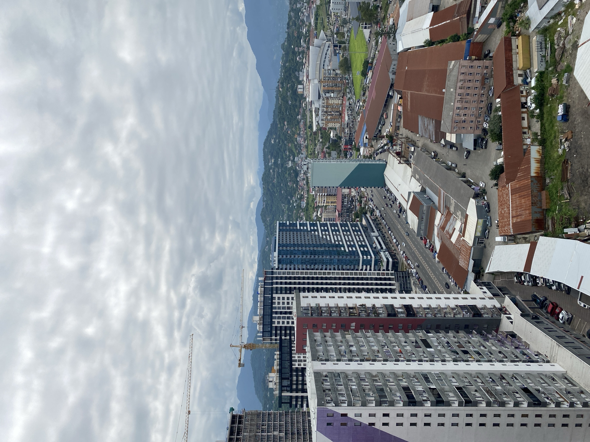 This is an aerial view of a city, likely Batumi, Georgia, on an overcast day. The scene is dominated by high-rise buildings, construction cranes, and a densely packed urban landscape. In the foreground, several modern residential buildings are visible, characterized by their varied colors and architectural designs, some featuring balconies. Adjacent to these residential towers are lower-rise commercial or industrial buildings with corrugated metal roofs. A street lined with parked cars and ongoing traffic runs through the middle ground. Beyond the immediate urban area, a sports stadium or large venue with a distinctive curved roof is present. Further in the distance, rolling green hills covered with scattered houses ascend to form a mountain range under a cloudy sky. The overall impression is one of a developing urban area with a mix of residential, commercial, and infrastructure elements.