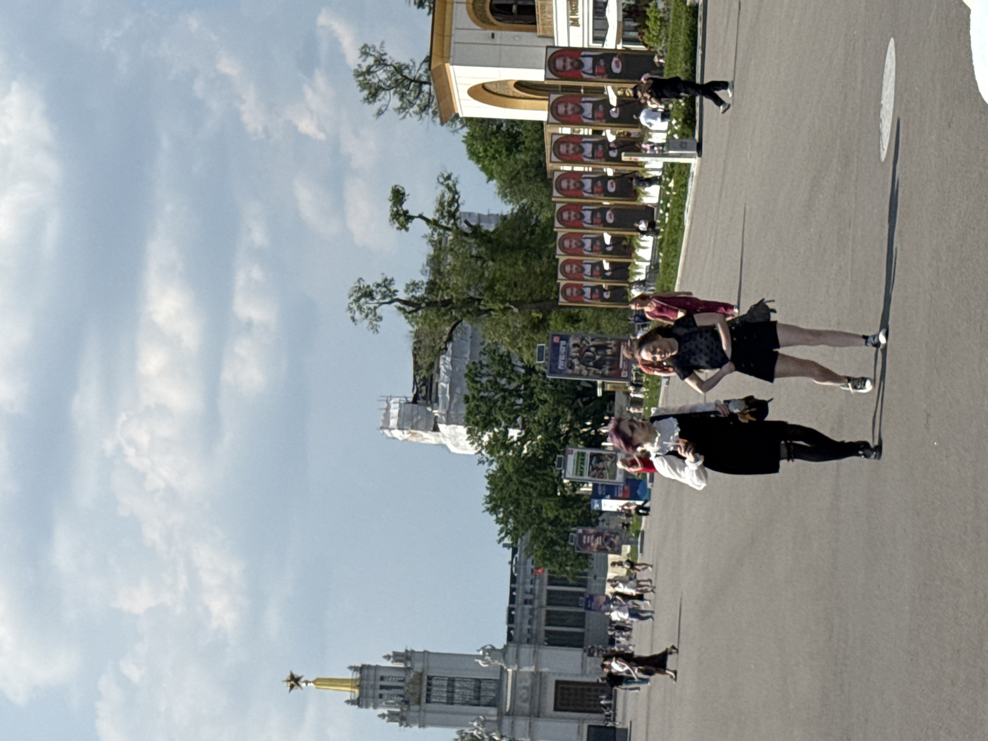 The image depicts a sunny day at the VDNKh (Exhibition of Achievements of National Economy) in Moscow, Russia. In the foreground, two young women walk side-by-side across a wide, paved area. The woman on the left wears a black dress, white collar, and dark thigh-high stockings, carrying a beverage and a small bag. The woman on the right is dressed in a black top and black shorts, with a purse slung over her shoulder. Several other people are visible in the mid-ground, walking and standing. In the background, prominent structures of VDNKh are visible, including the iconic Central Pavilion with its golden spire and star, and other exhibition pavilions. A row of large portraits featuring men in aprons is displayed, and trees dot the landscape. The sky is partly cloudy.