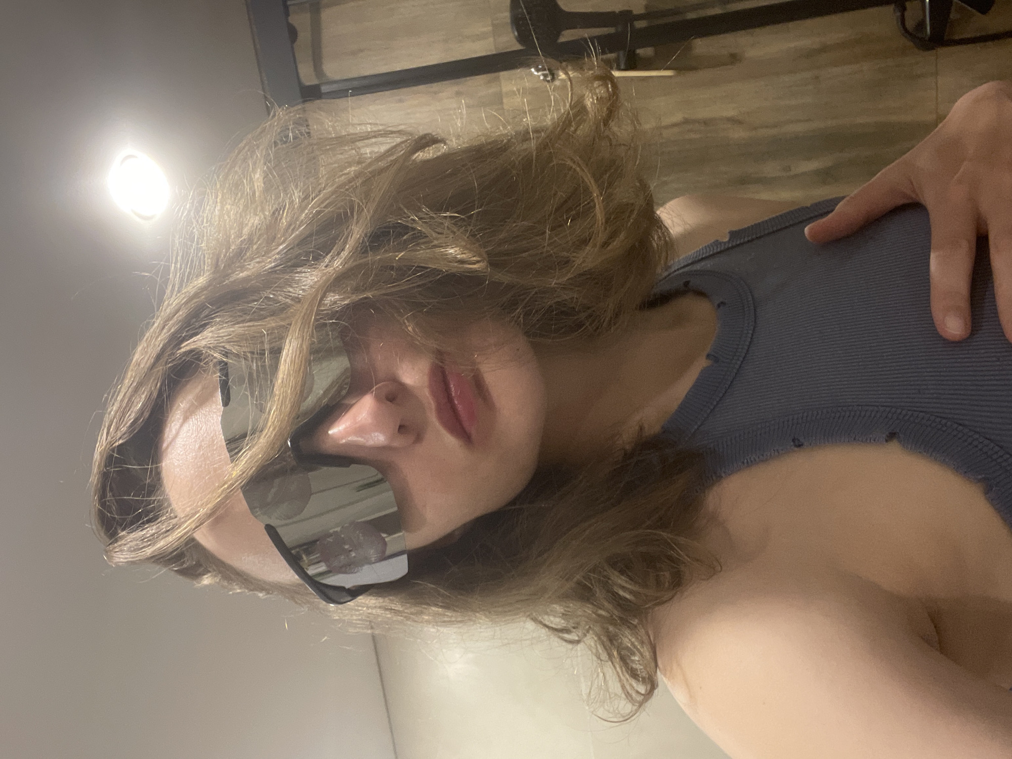 A woman with light brown, wavy hair is wearing reflective sunglasses and a ribbed, blue-grey tank top. She is indoors, with a light overhead. The background includes a wooden floor. Her hand is visible near her shoulder.
